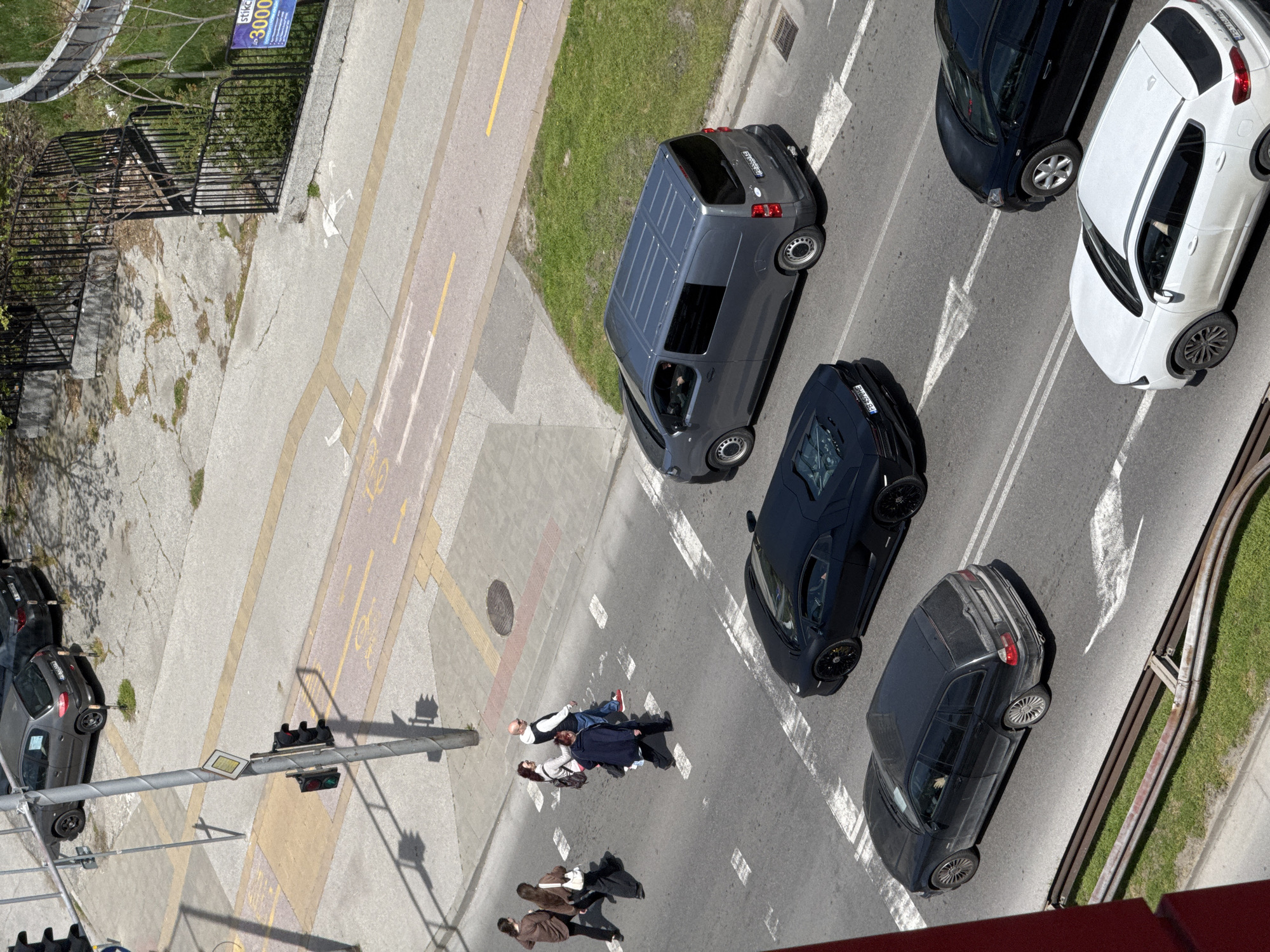 This high-angle image captures a bustling street scene in Plovdiv, Bulgaria, under bright, sunny daylight. The perspective looks down onto a multi-lane road, sidewalks, and a grassy strip, showing vehicles and pedestrians.
Several cars are visible on the asphalt road. A grey van (license plate PB 3393 CB) is positioned prominently, next to a sleek black sports car (PB 9099 HH), resembling a Lamborghini. Further along, a white hatchback (PB 7517 AB) and a dark grey sedan/station wagon (PB 0519 HH) are also present. Another dark grey car (PB 5262 HX) is partially visible on the bottom left. The vehicles appear to be parked or stopped, facing different directions.
A group of people is gathered on or near the road. In the foreground, a bald man in a white t-shirt and blue jacket stands near two other individuals. One person is in a dark jacket, in an unusual posture, seemingly lying or crouching very low on the ground next to another standing person. Further down the sidewalk, two more individuals are walking, one carrying a guitar case.
The road is marked with white arrows indicating traffic flow, double solid lines, and dashed lines. Alongside the main road, a distinct path with yellow and red-brown markings is visible, likely for cyclists or pedestrians, featuring white painted symbols on the ground. Traffic lights are mounted on a pole, showing both red and green signals. A black metal fence separates the street from an area of overgrown vegetation and trees. In the upper left corner, a blue sign partially displays the text "stikc" and "3000". Strong, clear shadows cast by objects and people indicate the time of day is likely midday or early afternoon.