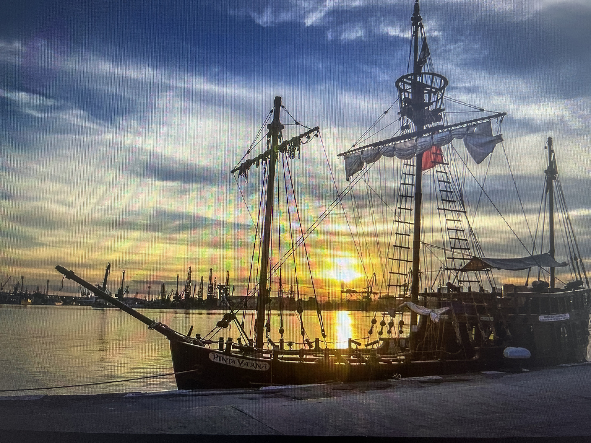 The image depicts the Pinta Varna, a replica of a historical sailing ship, docked in the port of Varna, Bulgaria. The ship is a prominent feature, with its masts, rigging, and partial sails silhouetted against a vibrant sunset. The sun's reflection creates a bright streak on the water's surface. In the background, the silhouettes of cranes and other port infrastructure are visible along the horizon. The foreground shows a concrete pier where the ship is moored. There are no visible people. The environment is characterized by the calm water of the port and the dramatic sky with clouds.