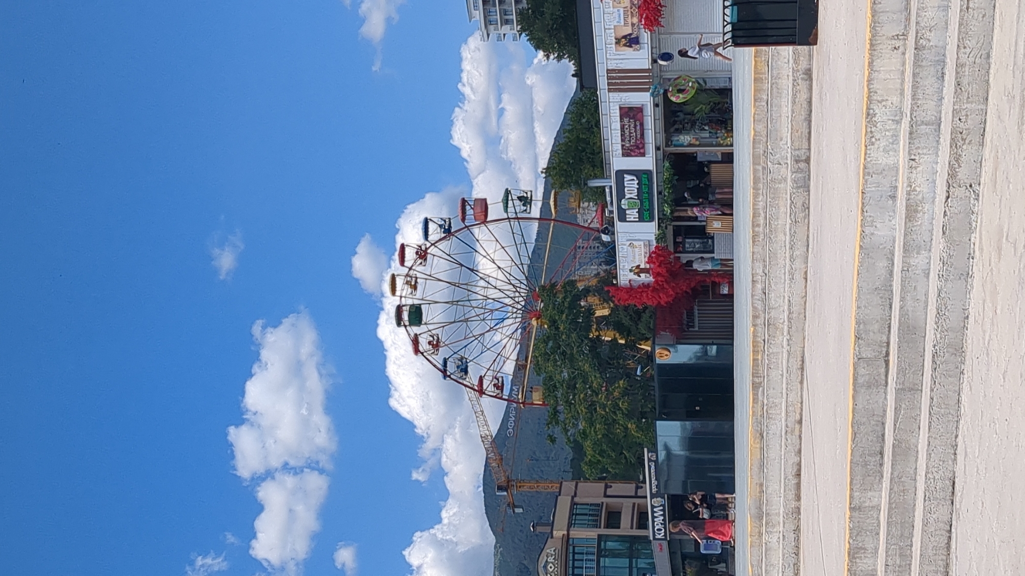 This image depicts a Ferris wheel with colorful cabins against a bright blue sky with scattered white clouds. The Ferris wheel is situated in an amusement area, with several small food kiosks or shops visible below it. One kiosk has a sign that reads "НА ХОДУ" (On the Go). A bright red, abstract sculpture is positioned near the kiosks. In the background, there are buildings, trees, a construction crane, and mountains. The foreground shows a concrete structure with multiple levels of steps. Several people are visible walking or standing near the kiosks and in the foreground. One woman in a red dress is walking towards the left side of the frame. The scene appears to be a sunny day in a city. The location is identified as Alushta, Ukraine.