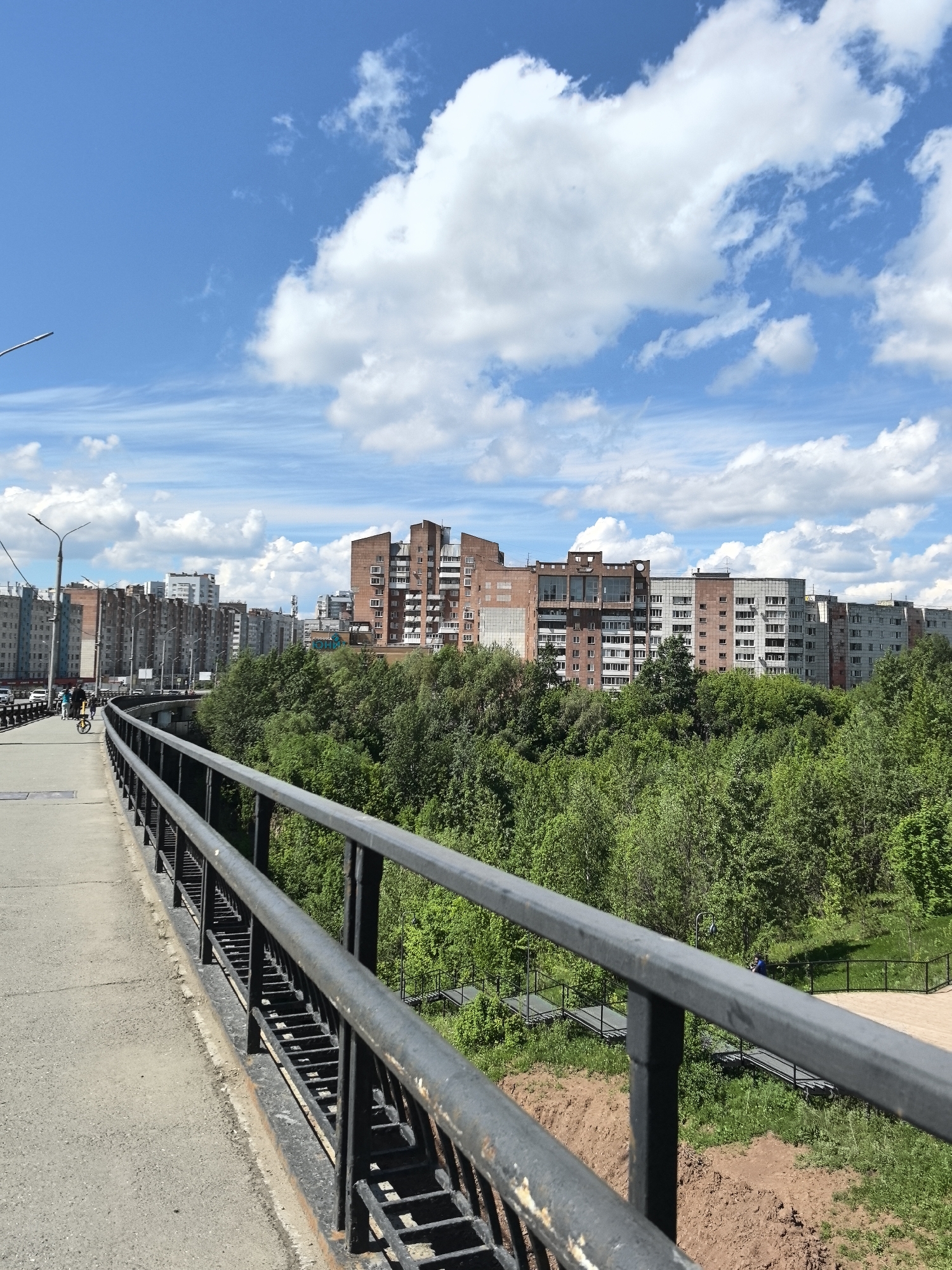 The image depicts a cityscape of Perm, Russia, taken from a high vantage point, likely a bridge. The foreground features a metal railing on a concrete walkway, presumably the edge of the bridge. A pedestrian and a cyclist are moving along the walkway in the distance. Beyond the railing, dense green foliage occupies the middle ground, obscuring some of the urban landscape. In the background, rows of buildings, varying in height and architectural style, stand against a bright blue sky scattered with cumulus clouds. A pedestrian bridge spans the ground beneath the bridge, adding a layer of complexity to the urban environment. The scene conveys a sense of urban life intertwined with natural elements.