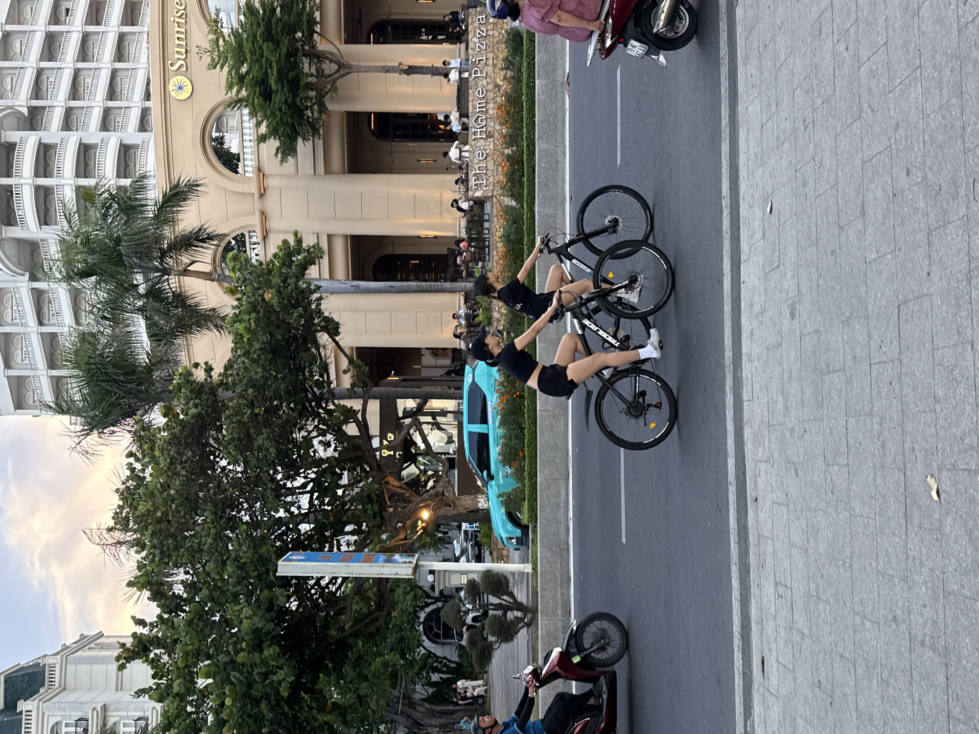 Two young women are riding bicycles down a street in Nha Trang Ward, Vietnam. The woman in front is wearing a black t-shirt and black shorts, with a black baseball cap. She is leaning forward and appears to be pedaling. The woman behind her is wearing a similar outfit: a black t-shirt, black shorts, and a black baseball cap. She is also leaning forward and seems to be pedaling. Both bicycles are black and have disk brakes.
The scene is a street with a sidewalk and buildings on the left. One building has "Sunrise" written on it in large letters, along with a circular logo. The architecture suggests it might be a hotel or a restaurant. There are trees and palm trees lining the street, and a turquoise car is parked on the side. Further down the street, there are more buildings, some with balconies and ornate details.
In the background, other people are visible, some seated at outdoor tables, suggesting a cafe or restaurant area. A red motorcycle is parked on the street, and a person wearing a blue shirt and a helmet is sitting on it. The lighting suggests it is late afternoon or early evening, with a soft glow in the sky and on the buildings. The weather appears to be pleasant, with some clouds visible in the sky. There is a sign on a pole with blue and white lettering, but the text is not clearly legible.