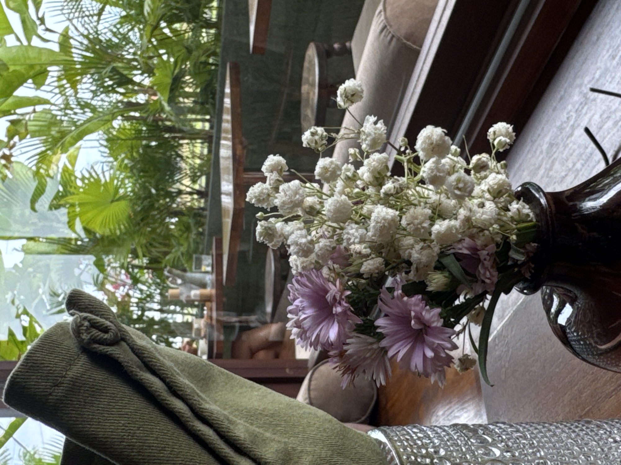 A close-up shot focuses on a bouquet of flowers in a dark brown vase placed on a wooden surface. The bouquet features delicate white baby's breath and several larger pinkish-purple aster flowers. To the left, a textured glass vase is partially visible. In the background, through a window, lush green foliage of tropical plants is evident, suggesting an indoor setting with an outdoor view. Several wooden tables and chairs are visible, hinting at a cafe or restaurant environment. The lighting is soft, contributing to a tranquil ambiance.