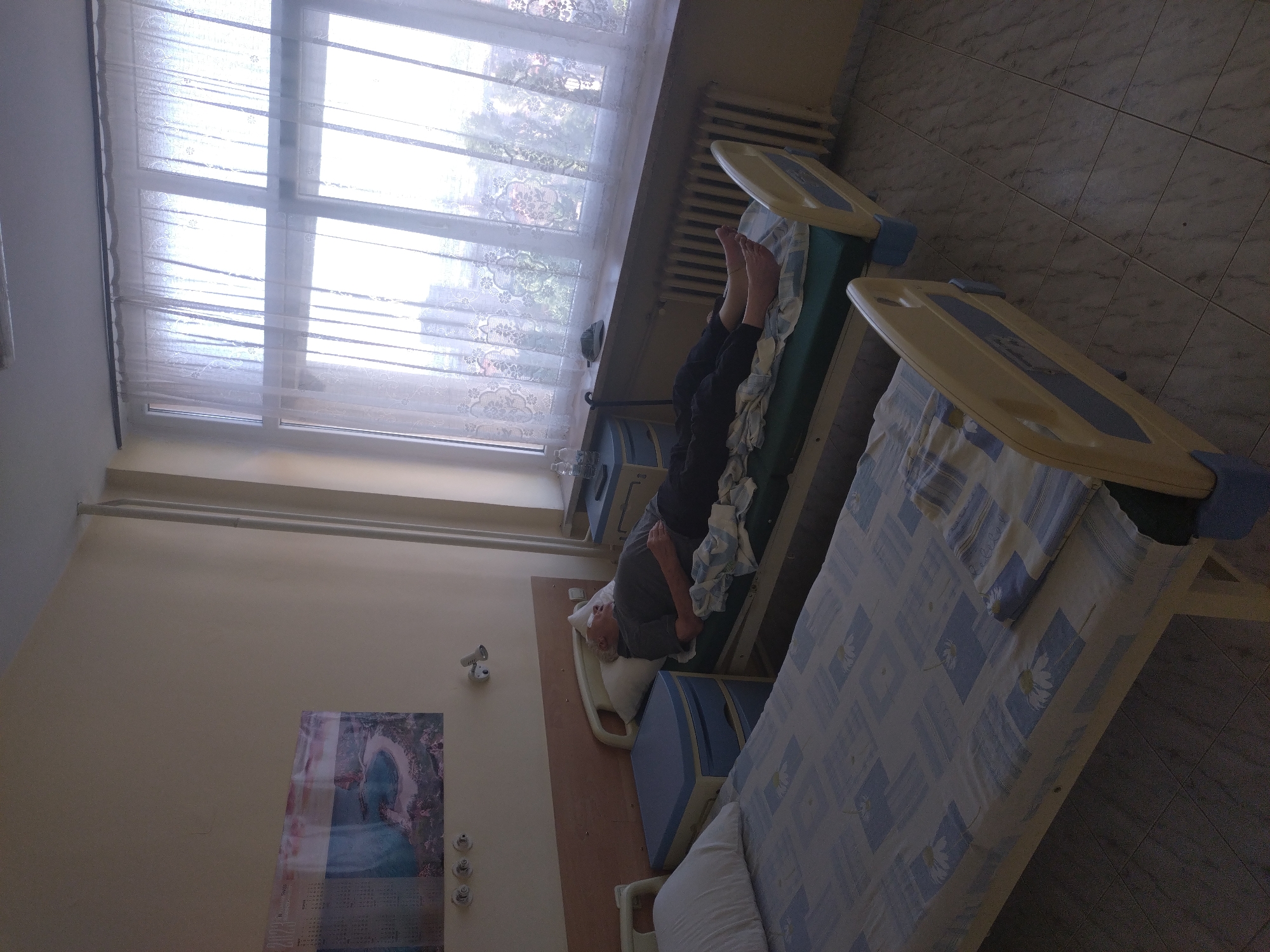 An elderly Caucasian male with light-colored hair is lying on a hospital bed in a dimly lit room. He is wearing a dark grey t-shirt and black pants. His eyes are closed, and he appears to be resting. The bed is covered with a blue and white patterned sheet and a white pillow. Next to this bed is another hospital bed with a blue and white patterned sheet and a white pillow. The room features a large window covered by sheer white curtains, revealing the faint outlines of buildings and trees outside. A radiator is visible beneath the window. A nightstand with a blue top is positioned between the two beds, and on it sits a bottle of water and a pair of glasses. A cane leans against the wall next to the nightstand. On the wall to the left, a framed picture of a coastal landscape hangs. The floor is tiled with a grey and white marbled pattern. The lighting in the room is subdued, with light coming primarily from the window. The scene depicts a hospital room setting.
