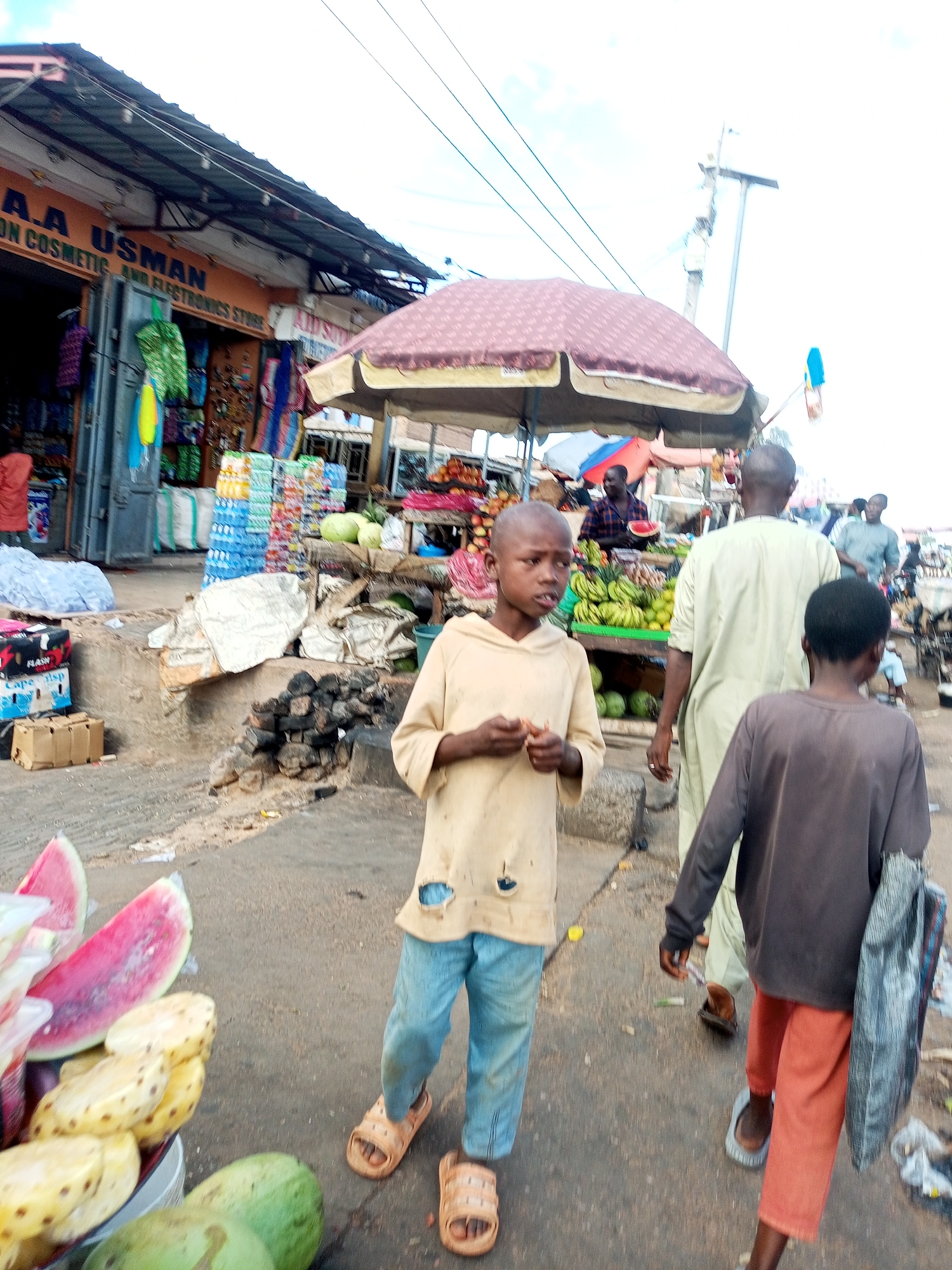 In Tempure, Nigeria, a bustling outdoor market scene unfolds. A young boy, wearing a light brown hooded shirt with ripped holes and blue jeans, stands in the foreground, holding small objects in his hands. To his right, another boy in a dark grey long-sleeved shirt and orange pants walks away from the camera. Behind them, a man in a light green robe also walks away, with other market-goers and stalls visible in the background. A stall displays an abundance of fruits, including watermelons and pineapples, and other items like bottled drinks and packaged goods. The environment appears to be a street with a paved surface, lined with shops and temporary market setups. Overhead, power lines crisscross the sky, and a utility pole is prominent. The overall atmosphere suggests a typical day in a lively Nigerian marketplace.