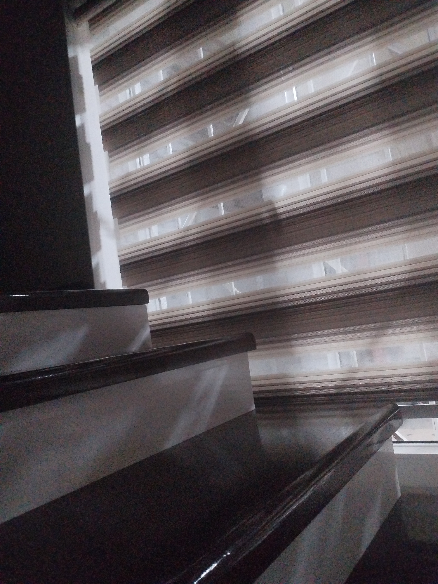A partially visible indoor staircase is shown in the foreground, with the steps covered in a dark, possibly varnished wood. The staircase railing is also dark. In the background, a window is covered by horizontal blinds with alternating opaque and sheer stripes, creating a patterned effect. The blinds are in shades of brown and beige. The overall scene appears to be an interior shot, possibly in a residential building. The location is described as Социализм, Kyrgyzstan.