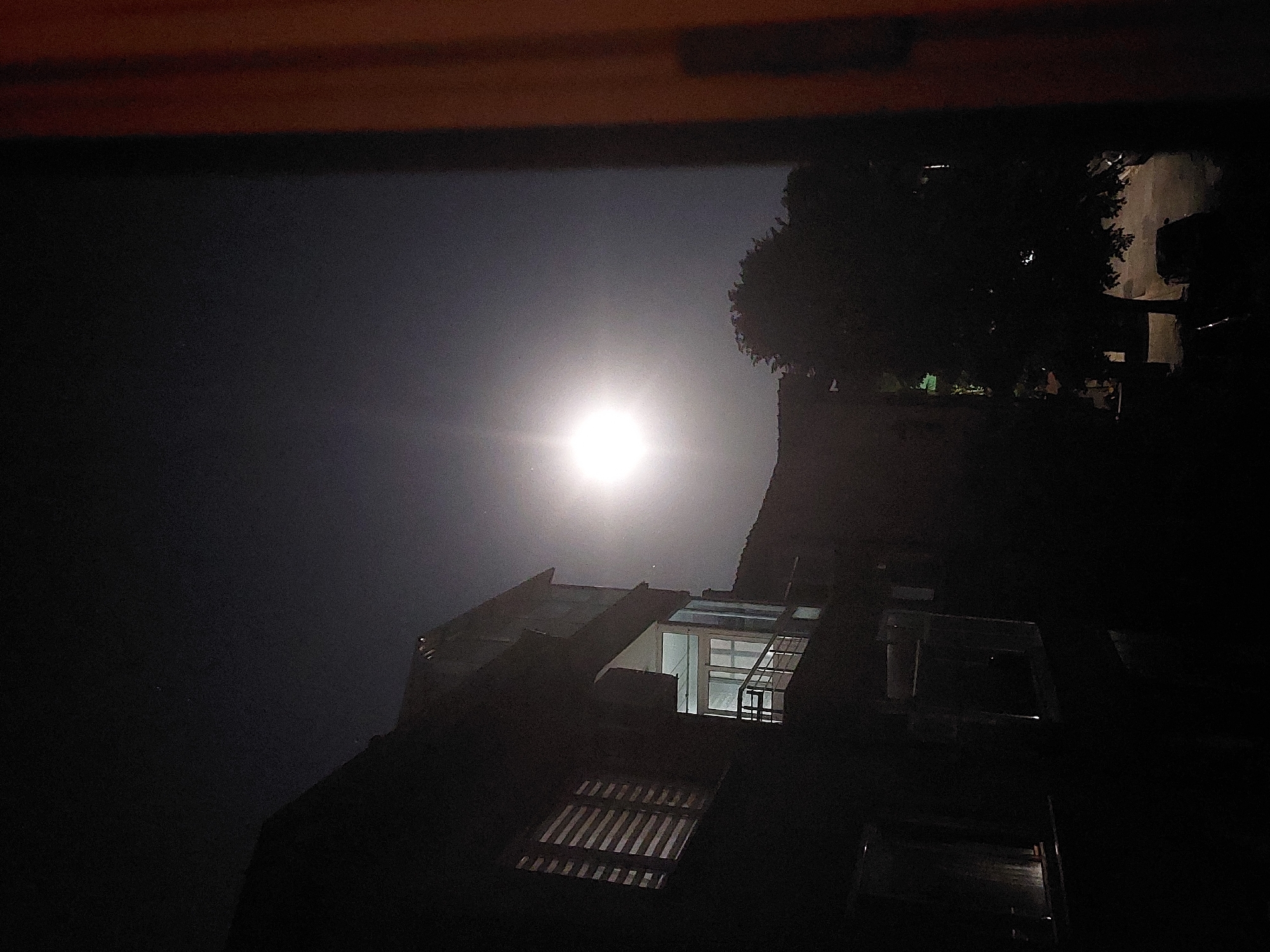 The image shows a nighttime scene of a building and sky. The bright moon is the primary light source. The building has a complex roof structure with visible skylights and windows. A tree partially obscures the building on the right side of the frame. The location is likely Tsarevo, Bulgaria, although this cannot be confirmed from the visual information alone. There are no people visible in the image. The environment is dark, suggesting a late hour. The overall scene depicts an urban setting at night under a bright moon.