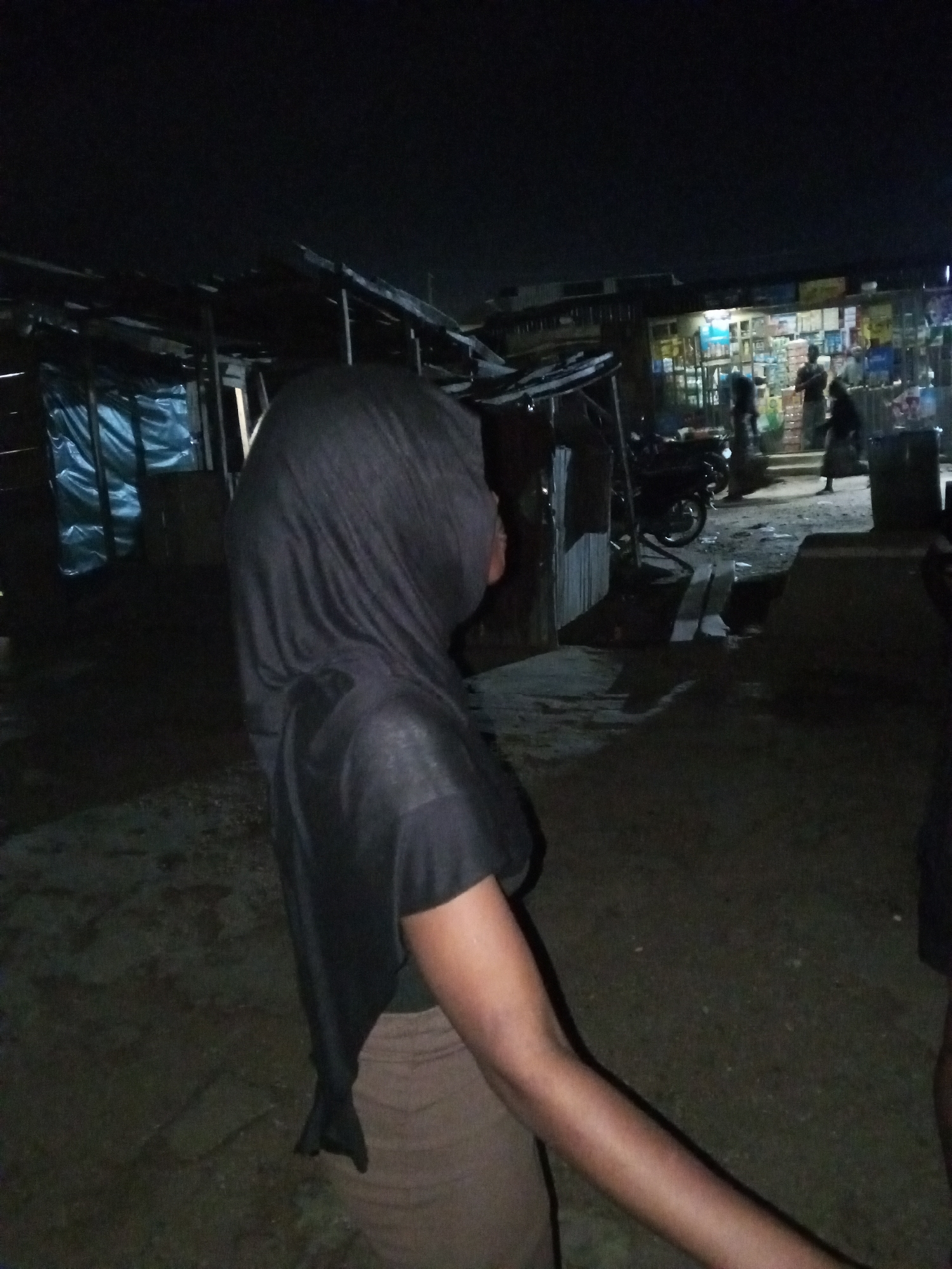 A medium shot shows a person from the side, wearing a black hijab and a black top, standing in an outdoor area at night. The person is facing to the right of the frame, with their head turned towards the background. The environment appears to be a market or street scene with stalls and a small shop in the background, illuminated by artificial lights. A motorcycle is parked in the middle ground to the left, and several people are visible in the background, including some standing near the shop. The ground is uneven and appears to be dirt or gravel. The lighting is dim, creating shadows and a somewhat grainy image quality. The location is described as being in the city of Lokoja, Nigeria.