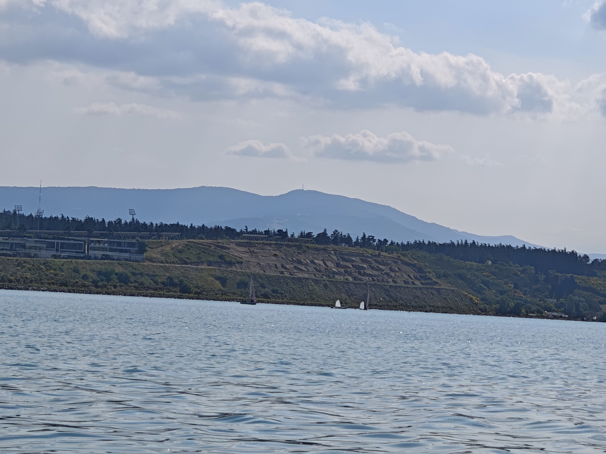 This is a wide, scenic shot of a body of water with sailboats in the distance and a lush, green hillside with buildings and trees in the background. The sky is partly cloudy, with puffy white clouds scattered across a light blue expanse. The time of day appears to be daytime, possibly late morning or early afternoon, given the brightness of the light and the shadows.
The foreground is dominated by the rippling surface of the water, which reflects the sky and creates a shimmering effect. Several small sailboats can be seen on the water, indicating a leisurely or recreational activity. They are small dots against the vastness of the water.
The middle ground features a terraced hillside covered in greenery, with a mix of trees and shrubs. On the upper part of the hillside, there are some modern-looking buildings, possibly residential or commercial, and some stadium lights are visible further to the left. The hillside appears to be a significant feature of the landscape, with a man-made terraced structure adding to its visual interest.
In the background, rolling hills fade into a hazy blue silhouette, suggesting a distant mountain range or a continuation of the landscape. A tall tower, possibly a communication tower, is visible on one of the higher peaks in the distance. The overall impression is one of a tranquil, natural setting with human development integrated into the landscape. The location context of Tbilisi, Georgia, suggests this might be a view of the Tbilisi Sea, a reservoir located north of the city, known for its recreational activities. No text is visible in the image.