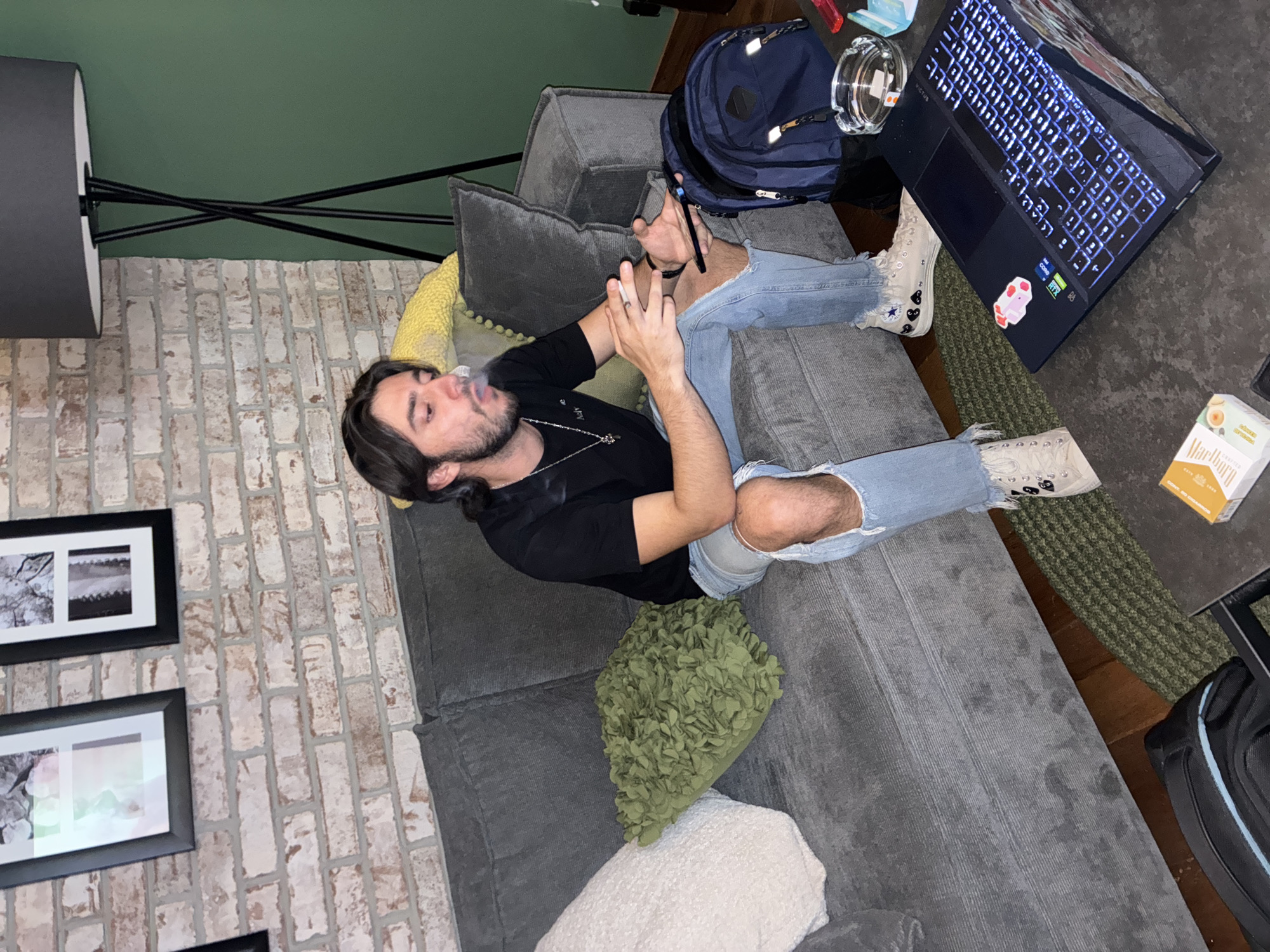 A man is lying on a gray sectional sofa. He is smoking. His legs are crossed, and he is wearing distressed jeans and sneakers with black hearts. His backpack is next to him. A laptop with a glowing keyboard is on the floor next to a clear ashtray, and a pack of Marlboro cigarettes are near the laptop. The room features a lamp, brick wall, and framed pictures. The location is not explicitly clear.
