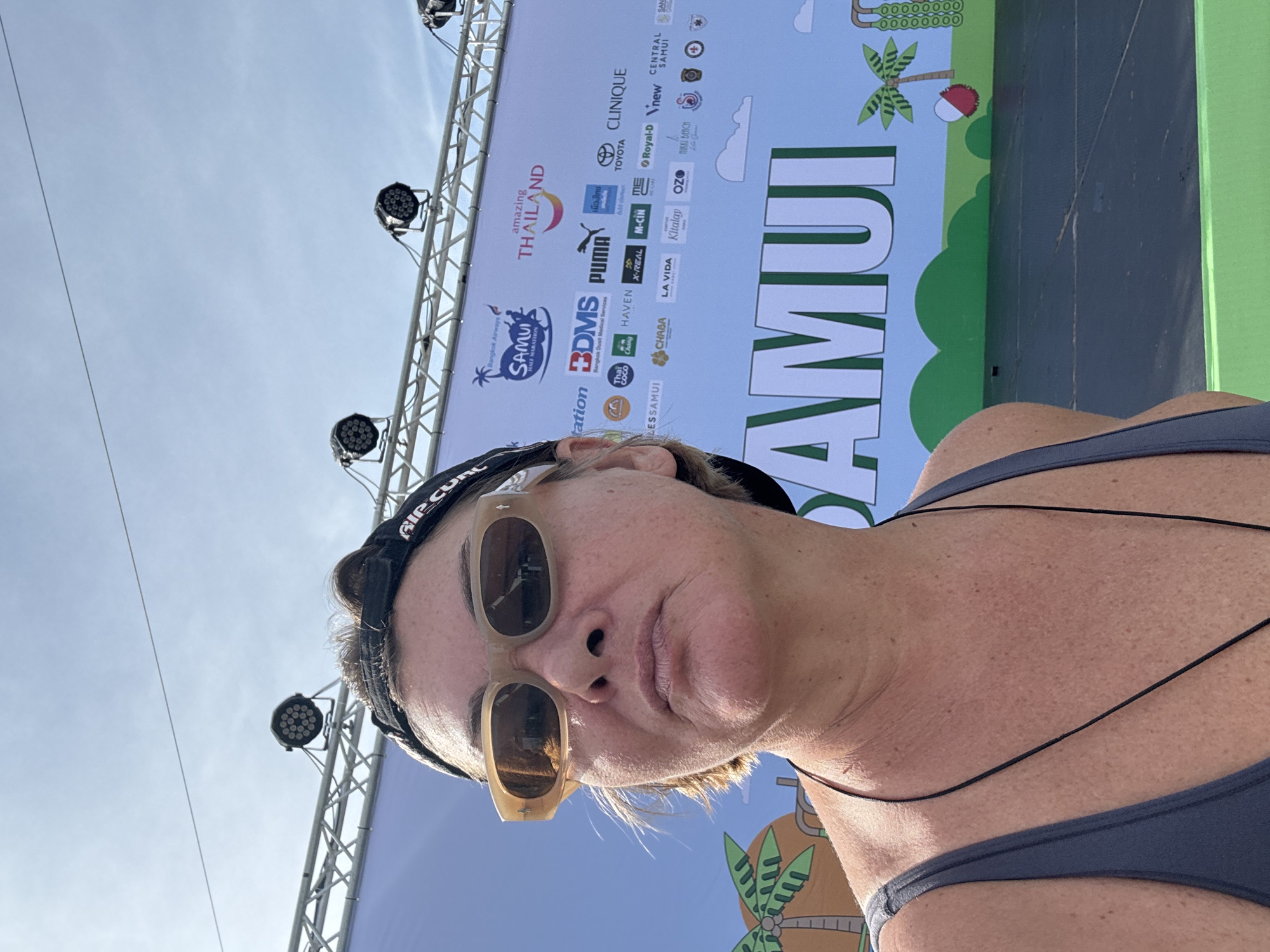 A woman with short brown hair, wearing sunglasses and a baseball cap, poses for a selfie in front of a large banner with text and logos. The banner appears to be for a "Samui Half Marathon" in "Amazing Thailand". The woman is wearing a grey tank top and a black necklace. The background shows a bright blue sky and stage lighting equipment. The overall scene suggests an outdoor event or festival.