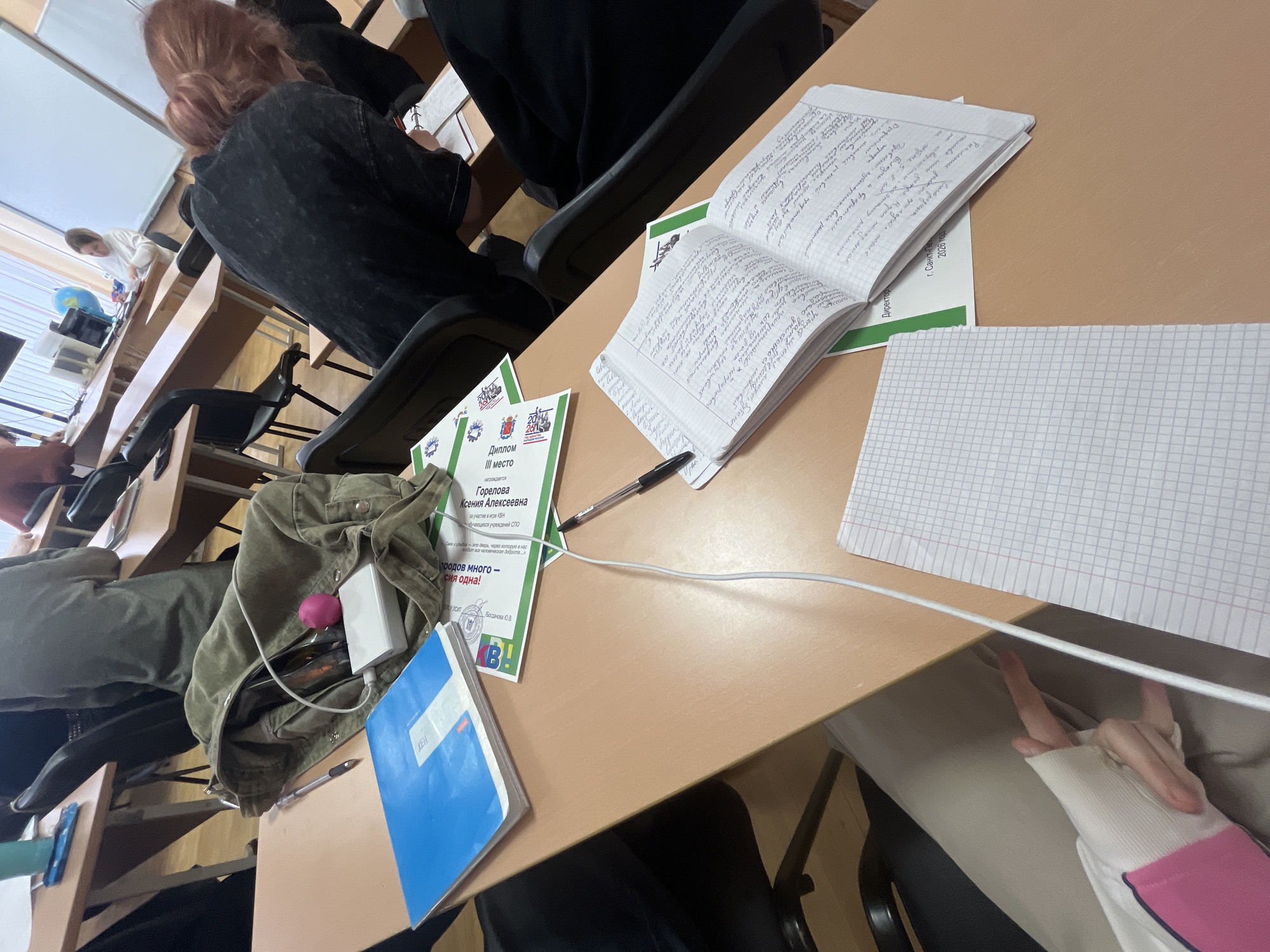 The image captures a scene in a classroom, likely in Saint Petersburg, Russia, based on the text on the diploma. The main subjects are the desks, notebooks, and people.
A diploma is lying on the desk, which reads "Diploma III place" and mentions "Gorelova Ksenia Alekseevna" being awarded for participation in a KVN game. The diploma also includes the phrase "prodov mnogo - siya odna!" and the name "Bogdanova Yu.V."
A notebook is open, with handwritten notes. Another notebook with a blue cover is present on the desk, along with a pen, a power bank, and a pink object inside an open bag.
People are seated at the desks, with a focus on a person with red hair. The classroom setting suggests an educational environment, possibly a university or college. There is a glimpse of another student at a desk by the window.