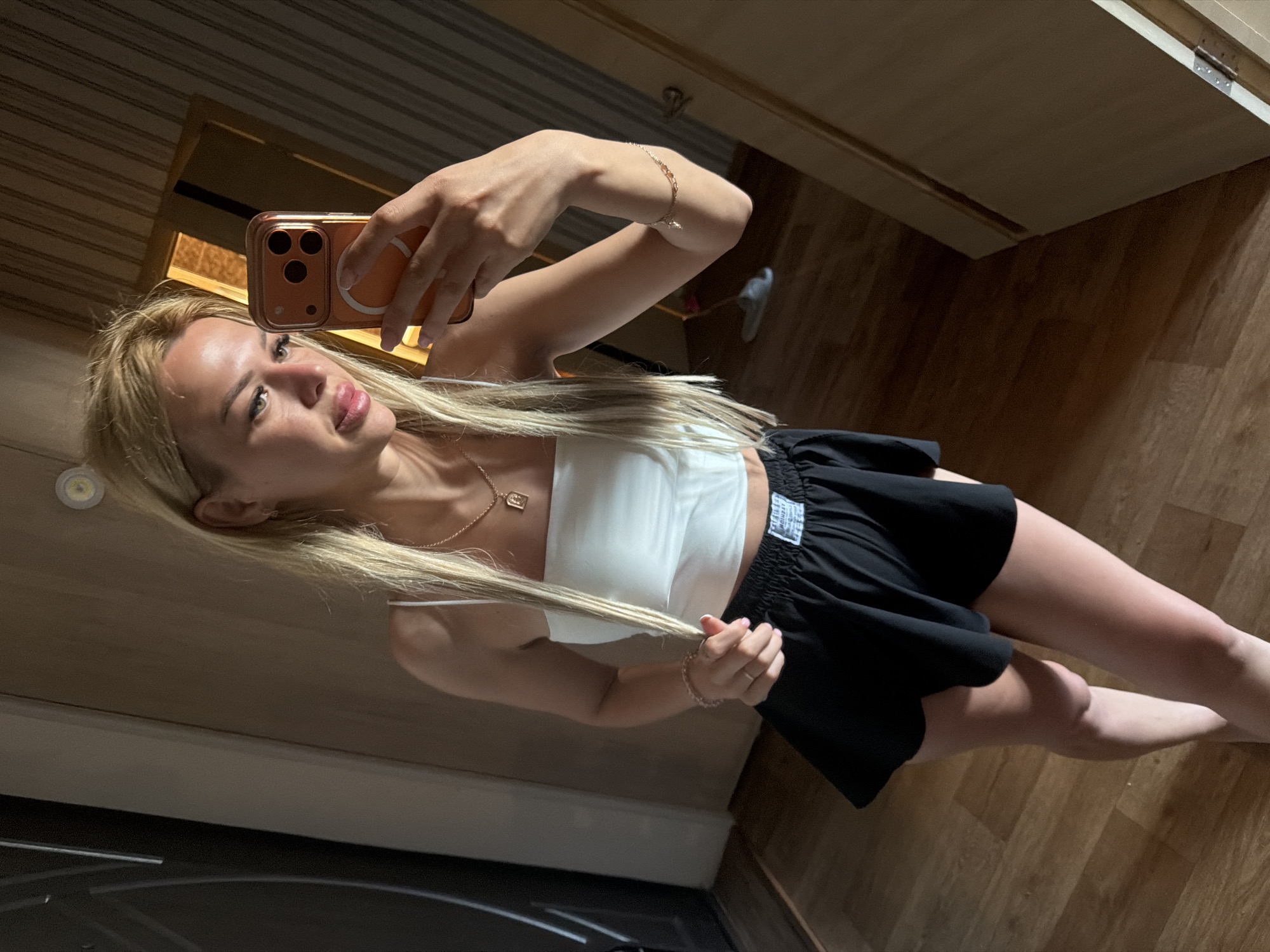 A young blonde woman is taking a mirror selfie in what appears to be a hallway or dressing area. She is wearing a white tube top and black shorts, and her hair is long and straight. She has a necklace and bracelets on. In the background, there is a door and a wooden floor. The lighting suggests it is daytime. The specific location in Kyiv, Ukraine cannot be determined from the image alone.