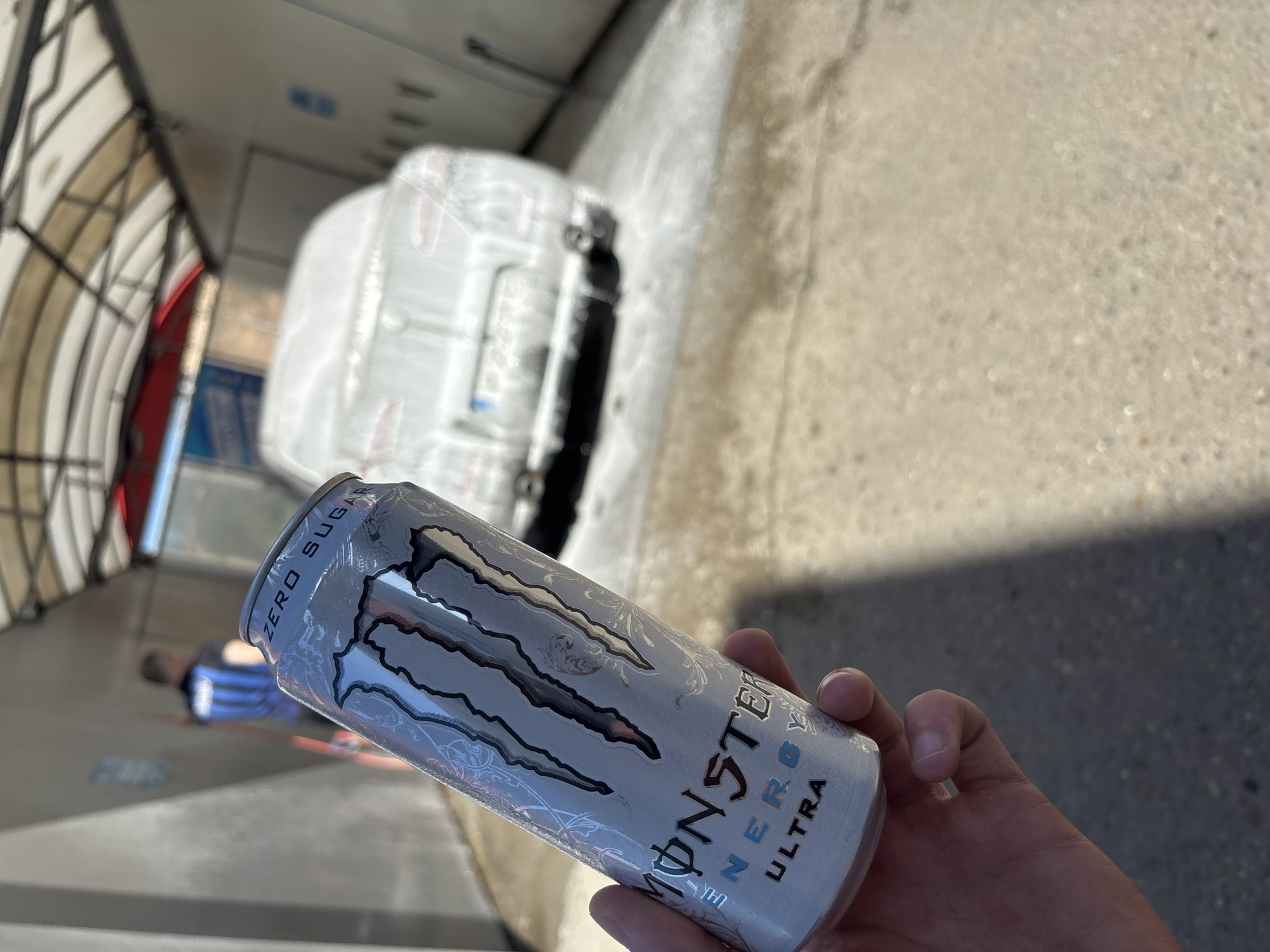 A hand holds a partially consumed can of Monster Energy Ultra drink in front of an open-roofed car wash in Ruse, Bulgaria. The car wash bay features a vehicle covered in sudsy white foam. The ground is light-colored concrete. Part of the car wash's framework is visible in the background. A person lies on the ground in the background wearing a blue and black striped shirt.