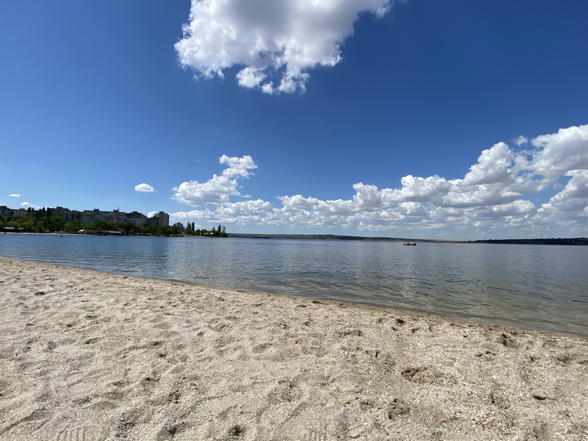 The image depicts a wide, sandy beach in the foreground with the sea stretching out to the horizon. The water is calm with gentle ripples. In the distance, a city skyline with buildings and trees can be seen along the left bank of the water. The sky is a clear blue, dotted with white, fluffy clouds. A small boat with two people is visible on the water in the middle distance. The overall scene is one of a serene, sunny day at the beach.