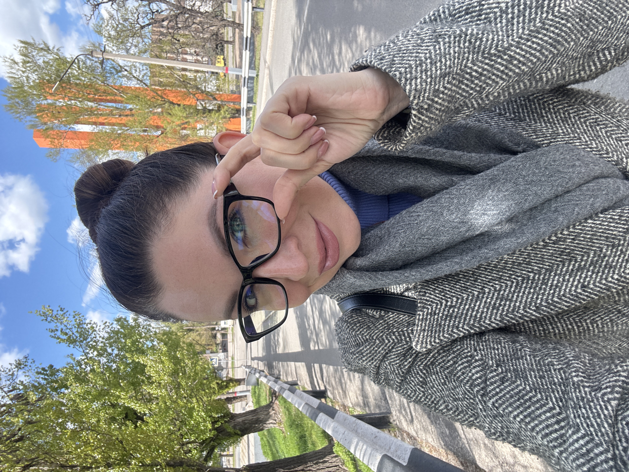 The image displays a waist-up self-portrait of a young woman outdoors in Kryvyi Rih, Ukraine. She has dark hair styled in a neat bun and wears black-framed glasses that reflect the bright sky and surrounding environment. Her eyes are a light color, and she has a calm, neutral expression. She is dressed in a grey coat with a prominent herringbone pattern, a matching grey scarf, and a blue top or sweater. Her right hand is raised near her face, possibly adjusting her glasses or shielding her eyes from the sun, revealing well-maintained nails with French tips.
The setting is an outdoor pathway or sidewalk, bordered by a black and white striped barrier that runs parallel to a road in the background. The scene is well-lit, indicating bright daylight under a blue sky with scattered white clouds. Trees line the path and road; some are lush with green foliage, while others have sparser branches, hinting at either different species or early spring. In the distance, partially obscured by trees, stand prominent tall orange rectangular structures, possibly a building or monument. No discernible text is visible in the image. The overall impression is one of casual observation in an urban or suburban environment.