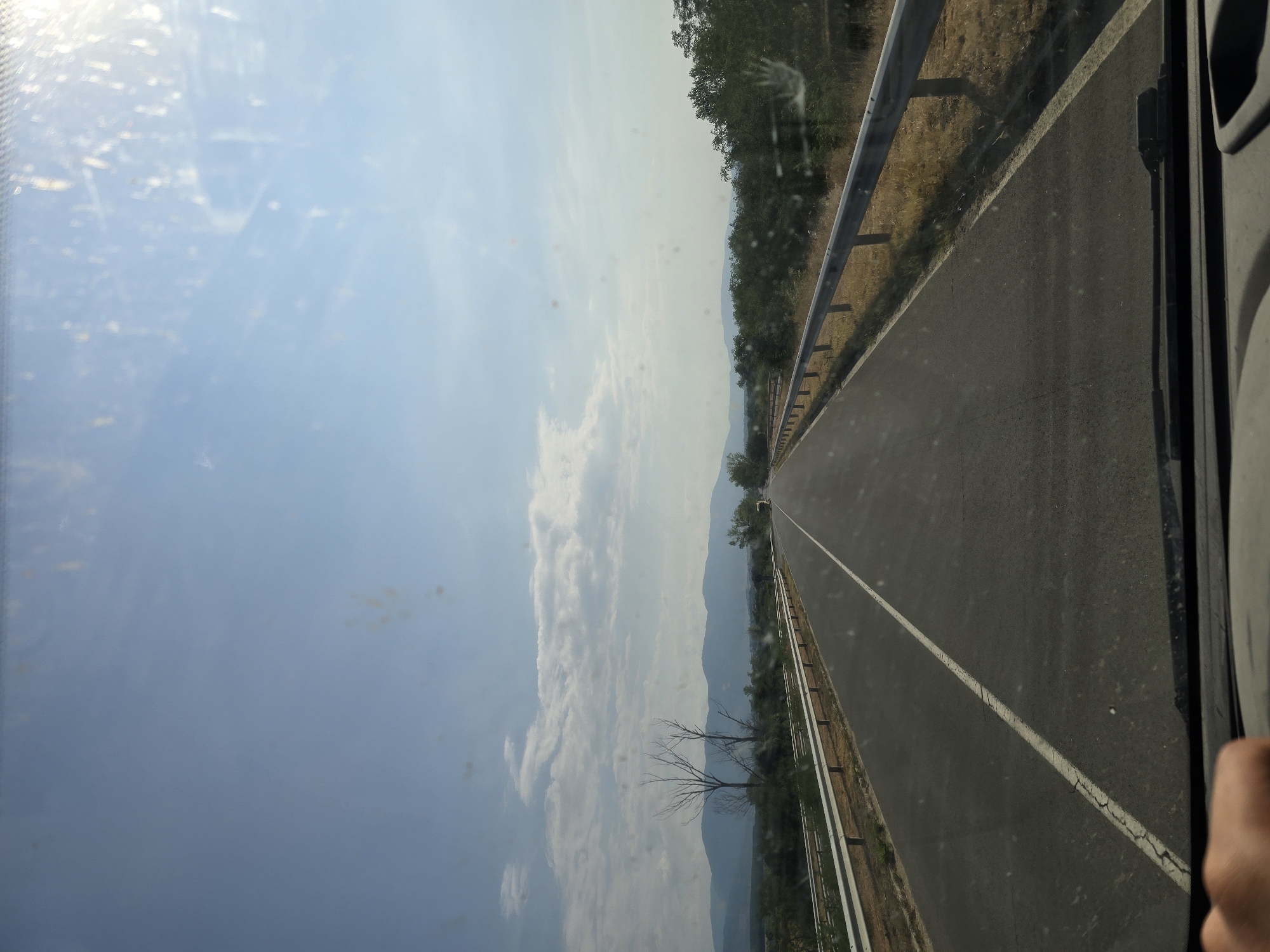 A first-person perspective shot from inside a vehicle on a clear day shows a paved road with a white line marking the lanes. The road stretches into the distance, flanked by guardrails on both sides. Beyond the road, dry grass and scattered trees line the right side, while the left side features more vegetation and a line of distant mountains under a blue sky with scattered white clouds. The image captures a highway scene, likely outside of Gurkovo, Bulgaria, given the context of the prompt. The windshield shows some dust and glare from the sun.