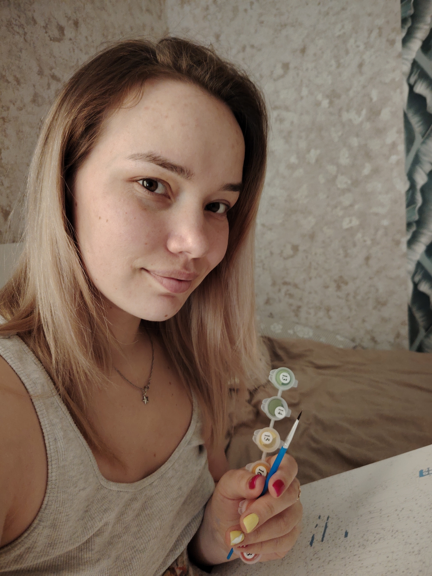 The image depicts a young woman indoors, likely in a private residence. She has fair skin, light brown hair with blonde highlights, and is wearing a light-colored tank top. She is holding a paint-by-numbers kit, including a set of small paint containers labeled with numbers and a paintbrush. Her fingernails are painted with alternating colors of yellow and red. Behind her is a wall with a textured, neutral-colored pattern, and part of a bed with a brown cover. The image appears to be taken in Марковский, Russia. The scene suggests she is engaged in a leisure activity, painting.