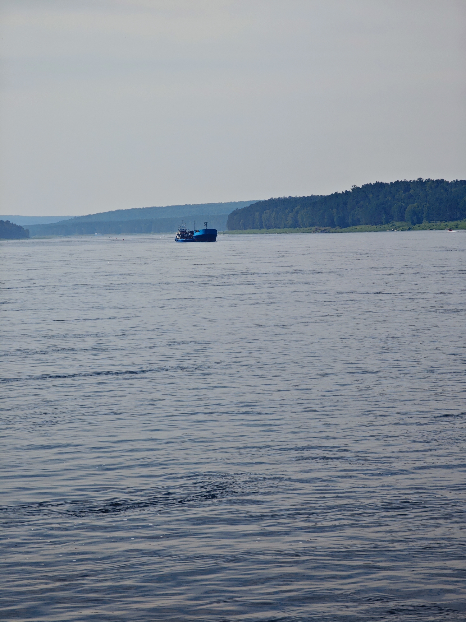 The image depicts a wide, calm river with gentle ripples reflecting the overcast sky. In the middle distance, a blue barge with several structures on its deck is moving across the water. The riverbanks are lined with dense green trees, forming a natural, forested landscape. In the far distance, layered hills fade into a hazy sky, suggesting a broad, open expanse. The scene is serene and natural, with no prominent urban structures visible. The location is identified as the city of Podporozh'e, Russia, which is situated on the Svir River, known for its navigable waterways and surrounding forests.