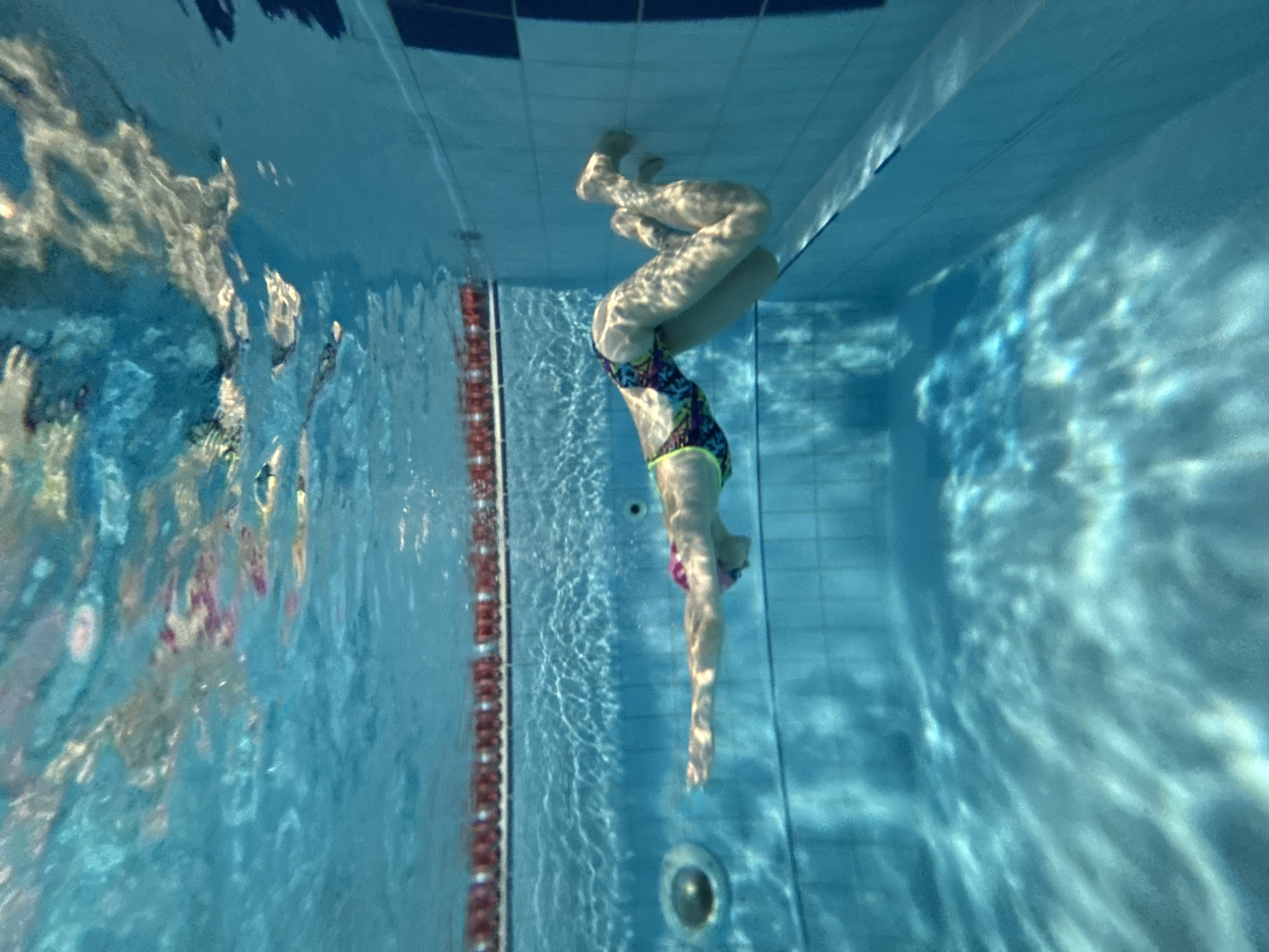 An underwater, full shot shows a person swimming in a swimming pool. The person is wearing a brightly colored swimsuit and a pink swim cap. Their arms are extended forward, and their legs are bent at the knees. The pool water is clear and blue, with light reflecting off the surface. The pool has tiled walls and a red lane marker at the top. The scene is located in the city of Chaikovsky, Russia.