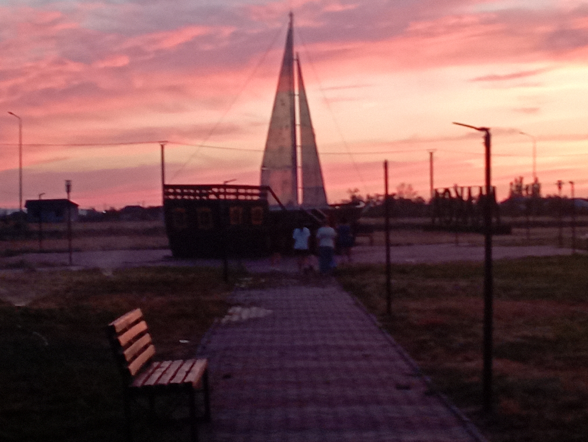 A photograph taken at dusk shows a surreal landscape dominated by a large, stylized sailboat structure. The sky is a vibrant gradient of pink and orange clouds, indicative of a sunset. In the foreground, a paved pathway leads towards the sailboat, with a wooden bench on the left side. Several indistinct figures are visible near the sailboat, appearing to be walking or standing. The background features a blurry silhouette of structures and trees, possibly in a park or open area within a city. The overall impression is one of an artistic installation set against a dramatic sky.