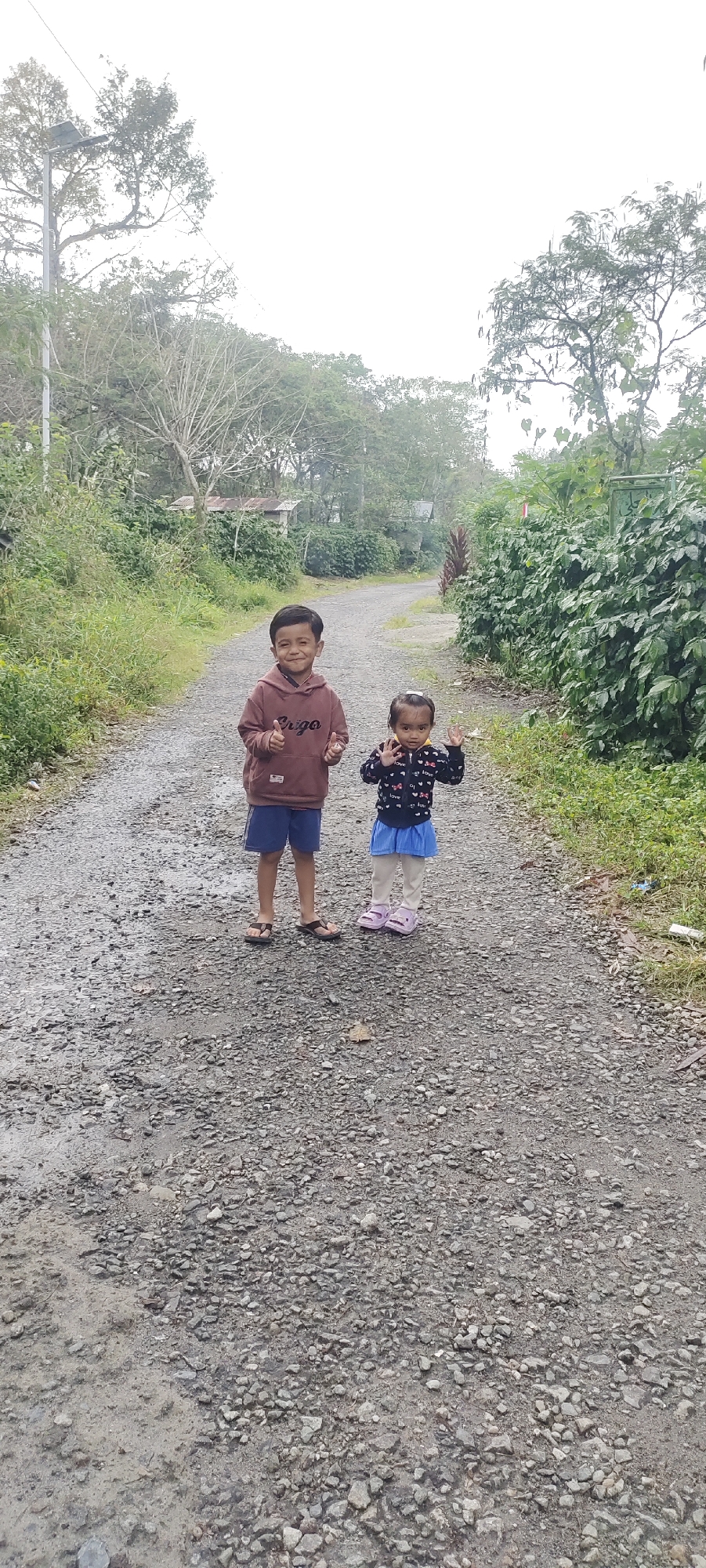 In Cintadamai, Indonesia, on a gravel road flanked by lush greenery and trees, a young boy and a little girl stand facing the camera. The boy, with dark hair and a broad smile, wears a maroon hoodie with the word "Cargo" printed on it and blue shorts. He gives a thumbs-up with his right hand. Beside him, the girl, with dark hair pulled back, wears a dark jacket adorned with heart and "love" motifs, and a blue skirt. She is holding her hands up, waving. The road appears damp, suggesting recent rain. In the background, a small structure with a corrugated roof is visible amongst the dense foliage, and a street lamp stands tall on the left side of the road. The overall atmosphere is one of casual outdoor activity in a rural setting.