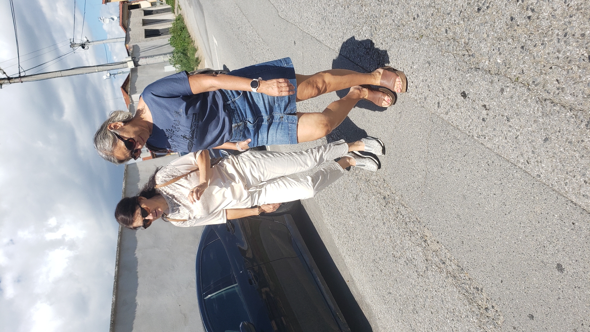 Two women are walking on a paved road with a slight incline. The woman on the left is wearing a cream-colored top and cream-colored pants, with silver sneakers. The woman on the right is wearing a navy blue t-shirt and a denim skirt, with brown wedge sandals. Both women are wearing sunglasses. A black car is parked on the left side of the road. In the background, there are buildings, a utility pole, and a cloudy sky. The scene is likely in Prešov, Slovakia, given the context.