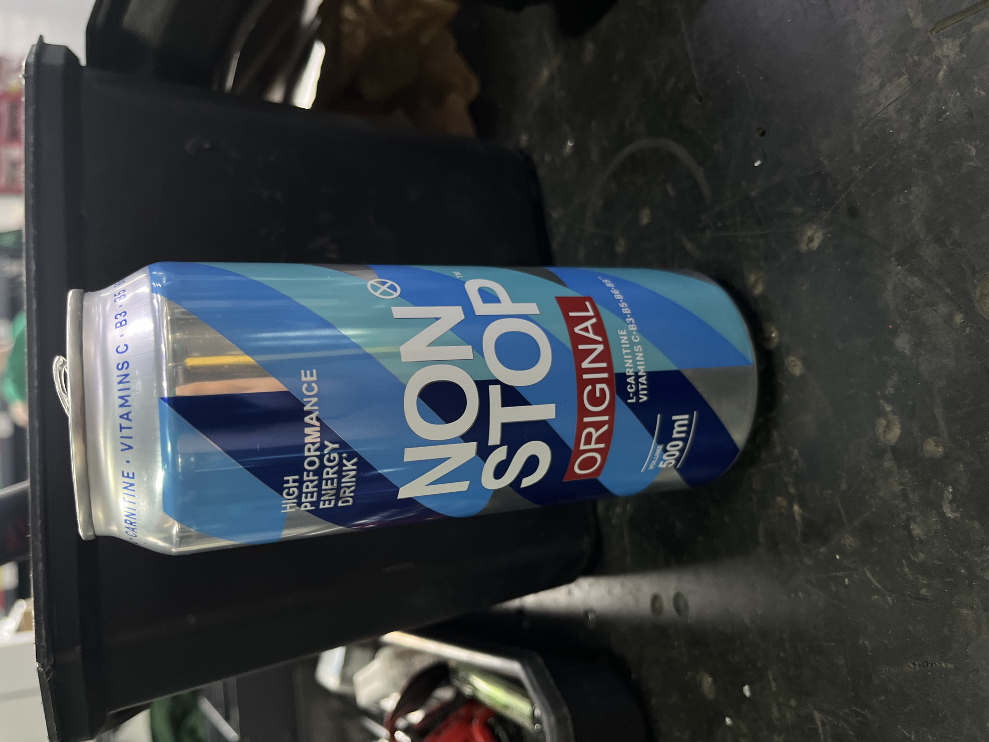 The image shows a close-up of a can of "NON STOP ORIGINAL" energy drink. The can is primarily blue with diagonal white and light blue stripes. The text "NON STOP" is prominently displayed in large white letters with a subtle shadow effect. Below this, "ORIGINAL" is written in red text.
On the left side of the can, smaller text reads "CARNITINE VITAMINS C B3 358" and "HIGH PERFORMANCE ENERGY DRINK". On the right side, further small text indicates "L-CARNITINE VITAMINS C B3 B5 B6 B9" and "VOLUME 500 ml".
The can appears to be placed on a dark, textured surface, possibly a table or workbench, with some blurred objects in the background, suggesting an indoor setting. The lighting is artificial, casting shadows and highlighting the metallic sheen of the can. The image is oriented such that the can is viewed from the side, tilted slightly upwards. There are no people visible in the frame. The overall impression is a product shot of an energy drink. The location context provided is Kyiv, Ukraine, but there are no visual cues in the image itself to confirm this.