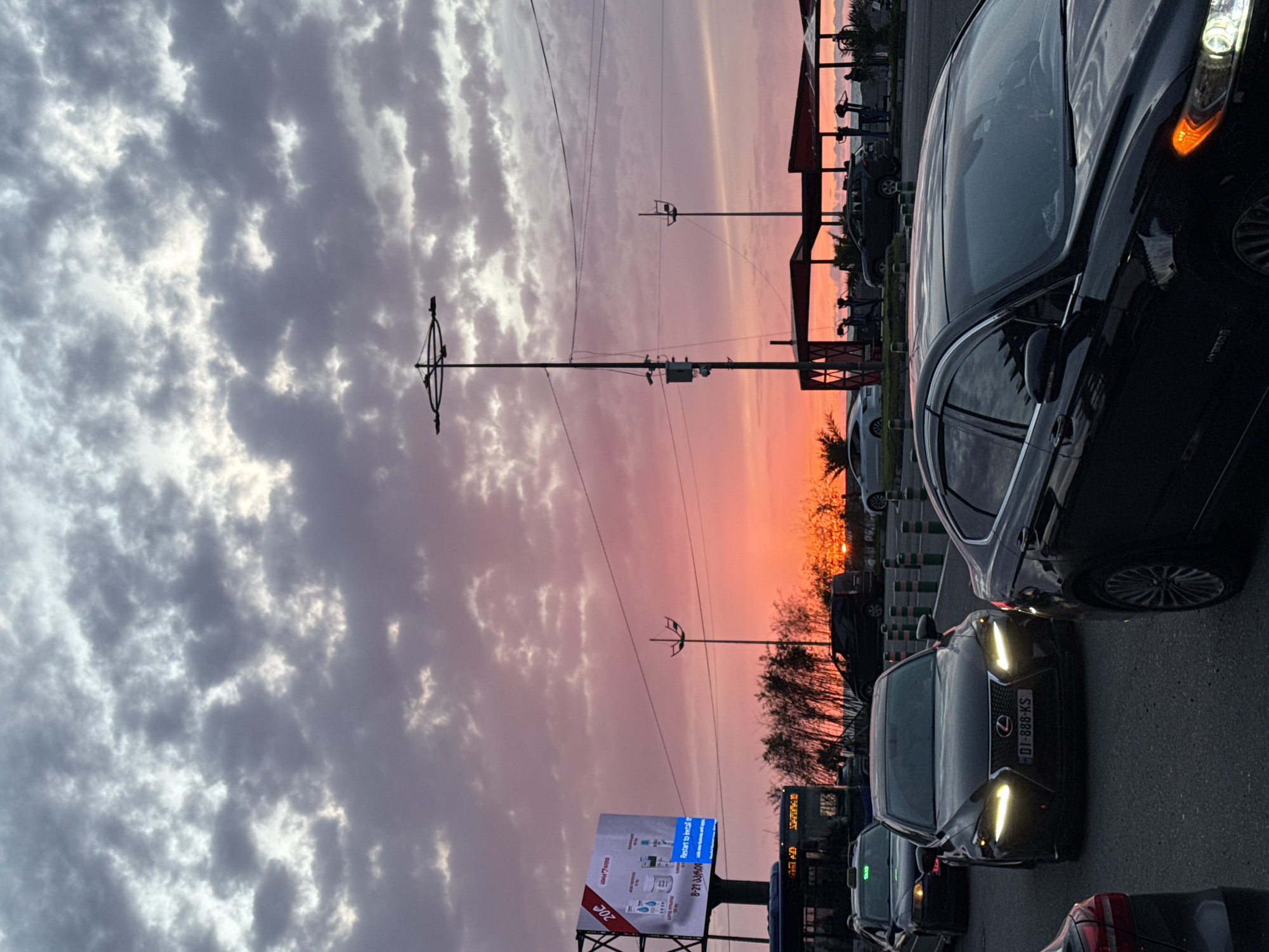 The image depicts a street scene in Batumi, Georgia, at sunset. The sky is a mix of grey clouds and vibrant pink and orange hues. Utility poles and wires traverse the sky. The foreground is filled with cars in traffic, including a black Lexus with the license plate "DI-888-KS". A billboard is visible on the left, advertising various products. Further down the road, trees and buildings are silhouetted against the sunset. People are vaguely visible in the distance under what appear to be awnings.