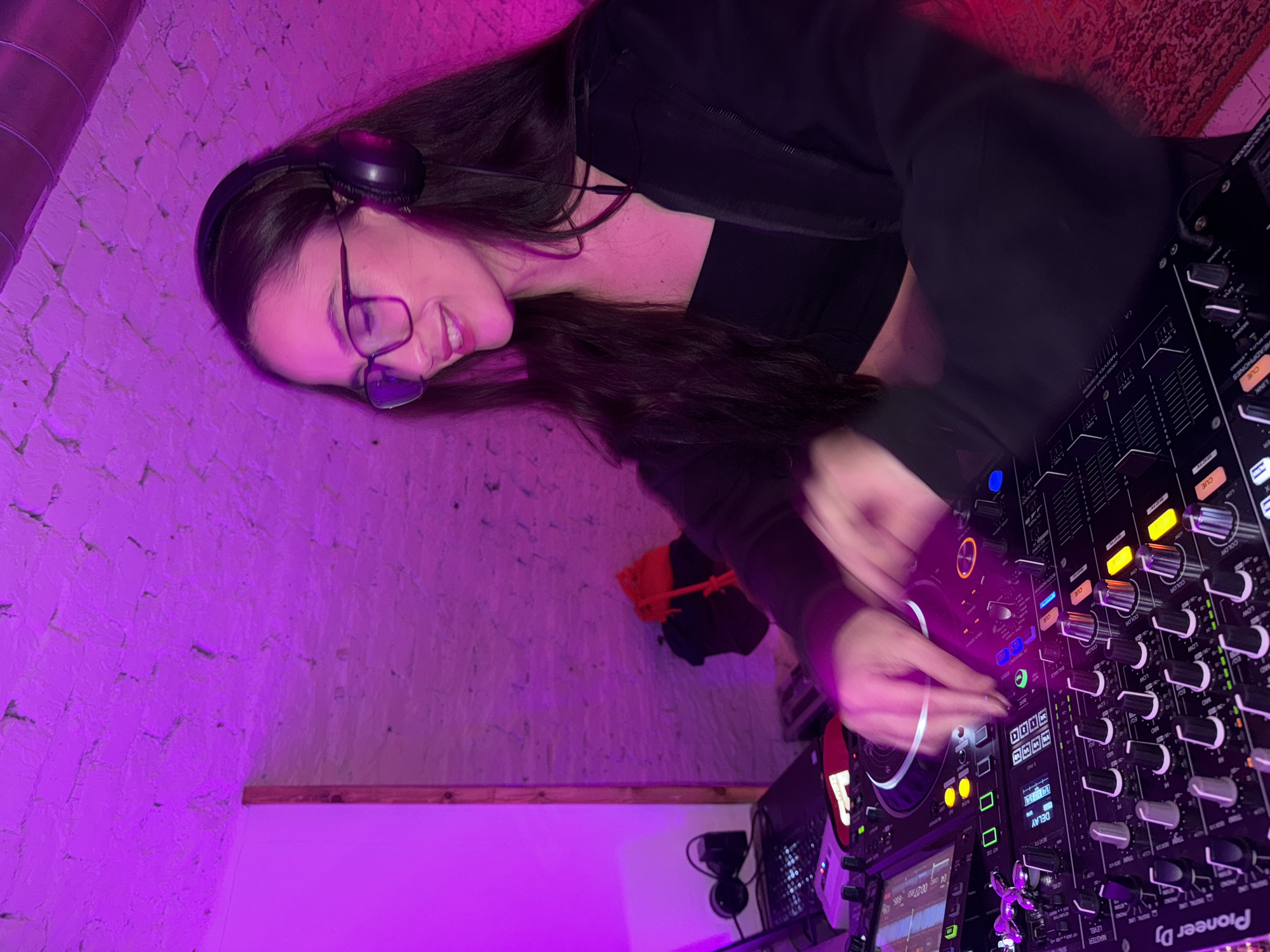 Here's a detailed description of the image:
**Overall Scene:** The image shows a woman DJing in what appears to be a club or a home studio setup. The lighting is dominated by a purple hue, possibly from LED lights or stage lighting.
**People:** A woman is the primary subject, wearing headphones, glasses, and a dark top. She's operating DJ equipment.
**Objects:** The main objects include professional DJ equipment: a Pioneer DJ mixer and CDJs. There's also exposed brick, a metallic pipe, a wooden beam, and additional equipment.
**Actions:** The woman is interacting with the DJ equipment, likely mixing tracks.
**Environment:** The setting seems to be an indoor space. The backdrop consists of a white brick wall and pipe.