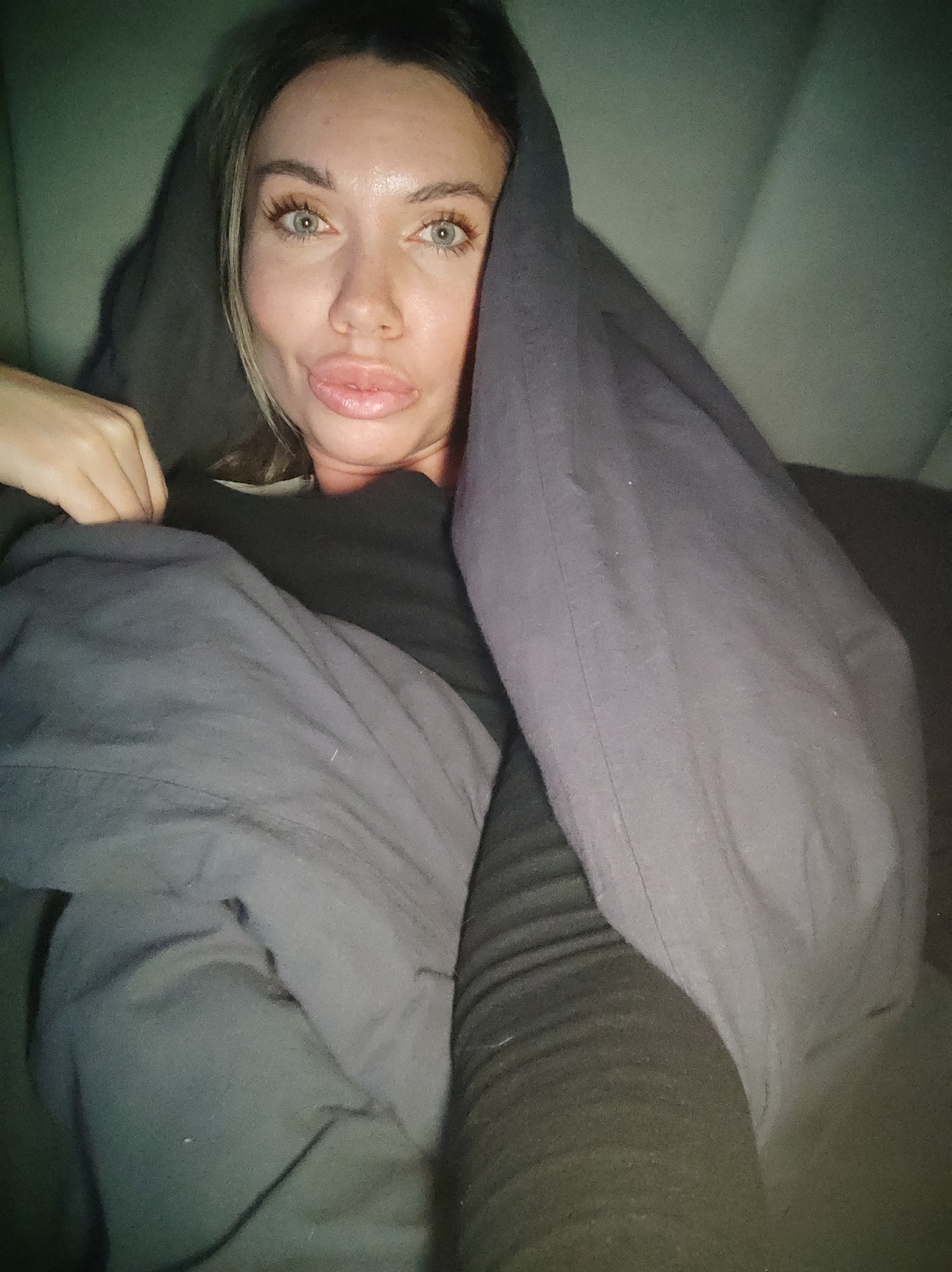 A woman lies in what appears to be a bed, covered partially by grey blankets. She is looking directly at the camera, with a blanket pulled over her head like a hood. The location is unclear and there is no evidence the image was taken in Votkinsk, Russia.
