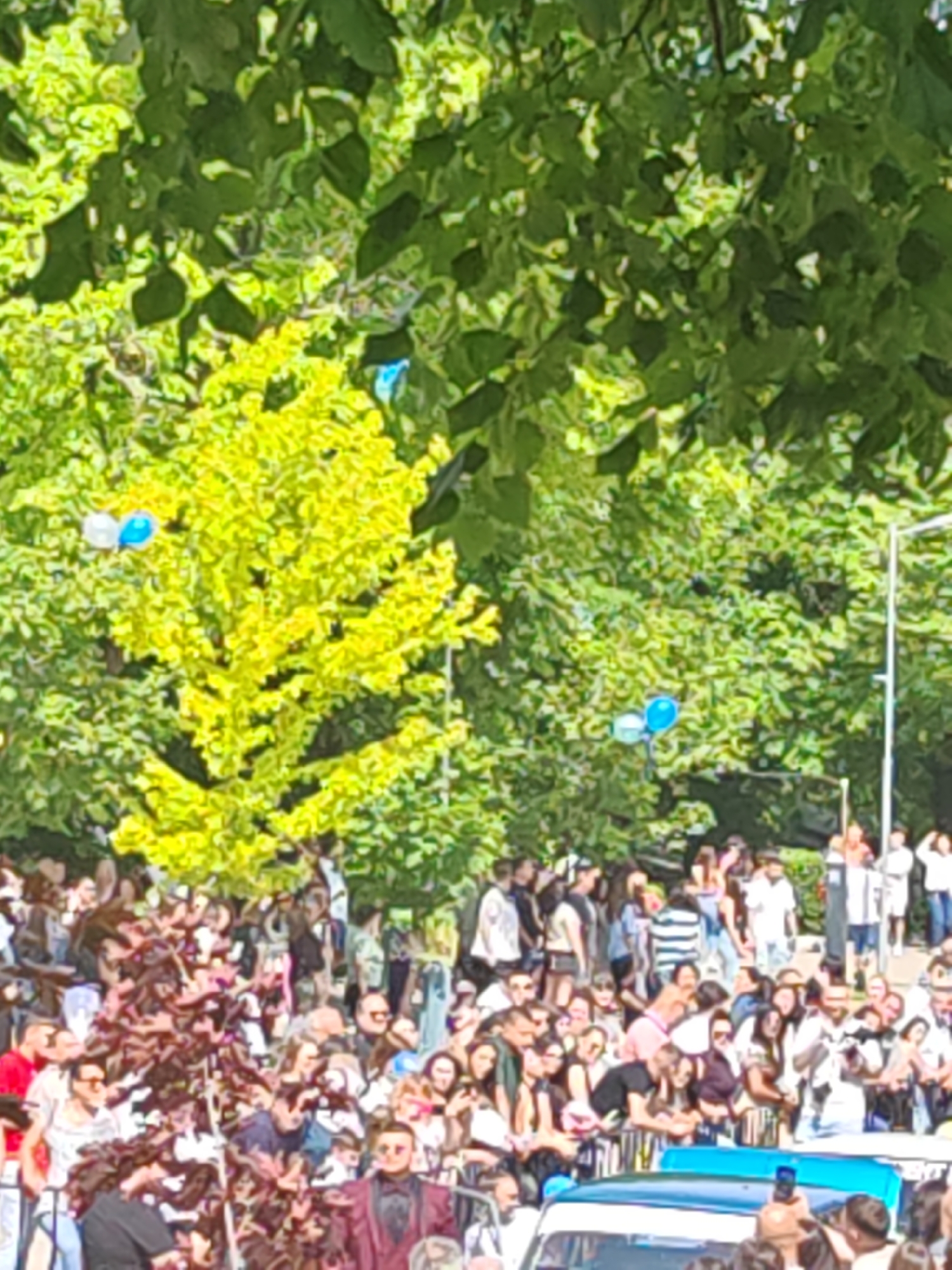 The image depicts a large crowd of people gathered outdoors, likely in a park or open urban space. Individuals of varying ages and appearances are present, many appearing to be spectators or attendees of an event. Trees provide a backdrop, and balloons are visible among the branches. Some objects suggest the possible presence of vehicles, which may be related to either authorities or the location of the event. While the image provides a glimpse of a gathering, it does not have enough detailed information to accurately describe this as happening in Shumen, Bulgaria.