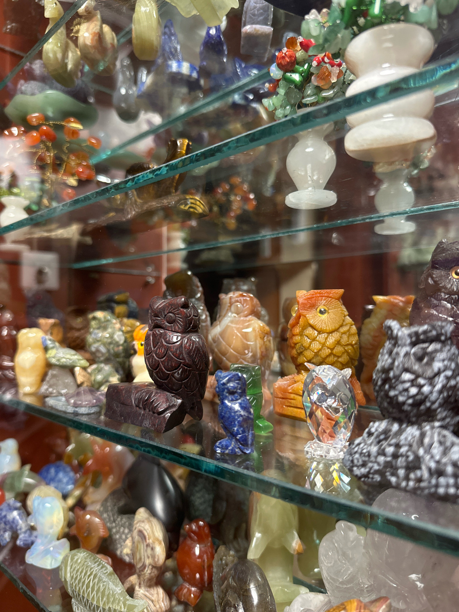 The image shows a close-up, slightly angled view of a glass display shelf filled with various carved stone figurines. The primary focus is on a collection of owl figurines in different colors and materials, including dark red, orange, and speckled black. There are also other animal carvings, such as fish, and decorative items like a beaded branch with red and orange elements and white vases. The lighting appears to be artificial, and reflections from the glass and surrounding objects are visible. The items are arranged on multiple glass shelves. The image does not contain any information about the city of Murmansk, Russia, or any people.