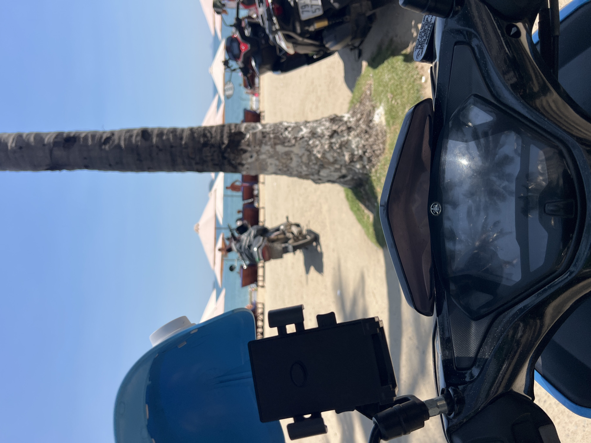 A close-up, ground-level shot shows a motorcycle parked near the beach in Bắc Nha Trang, Vietnam. The front of the scooter, with a blue body and black handlebars equipped with a phone holder, dominates the foreground. In the background, multiple motorcycles are parked on a sandy area beside a palm tree trunk. Beyond the motorcycles, the beach features white sun umbrellas and red lounge chairs facing the ocean. The sky is clear and blue.
