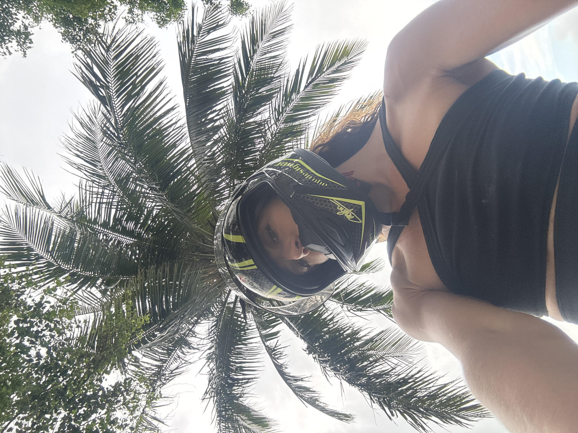 A low-angle, portrait-oriented photograph depicts a person wearing a motorcycle helmet, positioned against a backdrop of a palm tree and a cloudy sky.
The person is wearing a black tank top, and a black and yellow motorcycle helmet. Their face is partially visible through the helmet's visor.
The background consists of a tall palm tree with numerous fronds spreading out in a radial pattern. Above and around the palm tree, a greyish-white sky suggests an overcast day.
The location cannot be determined from the image.