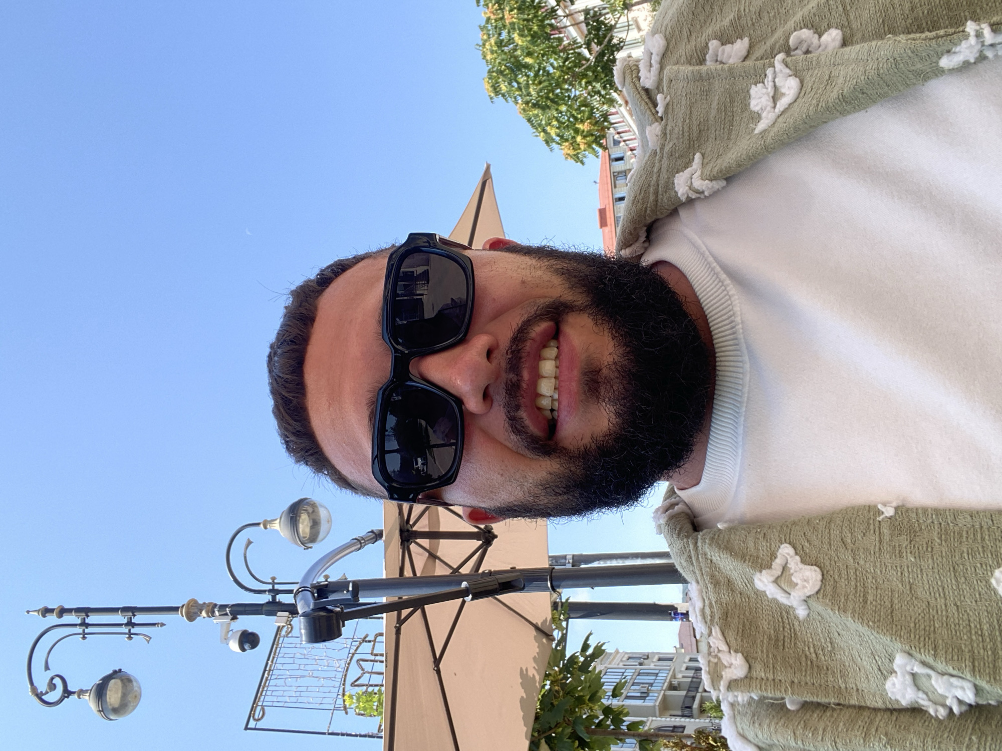 A medium shot shows a man with a beard and sunglasses looking directly at the camera. He is wearing a white sweatshirt and a green patterned jacket. Behind him, a lamppost with two lights and a security camera is visible, along with a beige umbrella and buildings. The sky is clear and blue. The scene is outdoors in the city of Derbent, Russia.