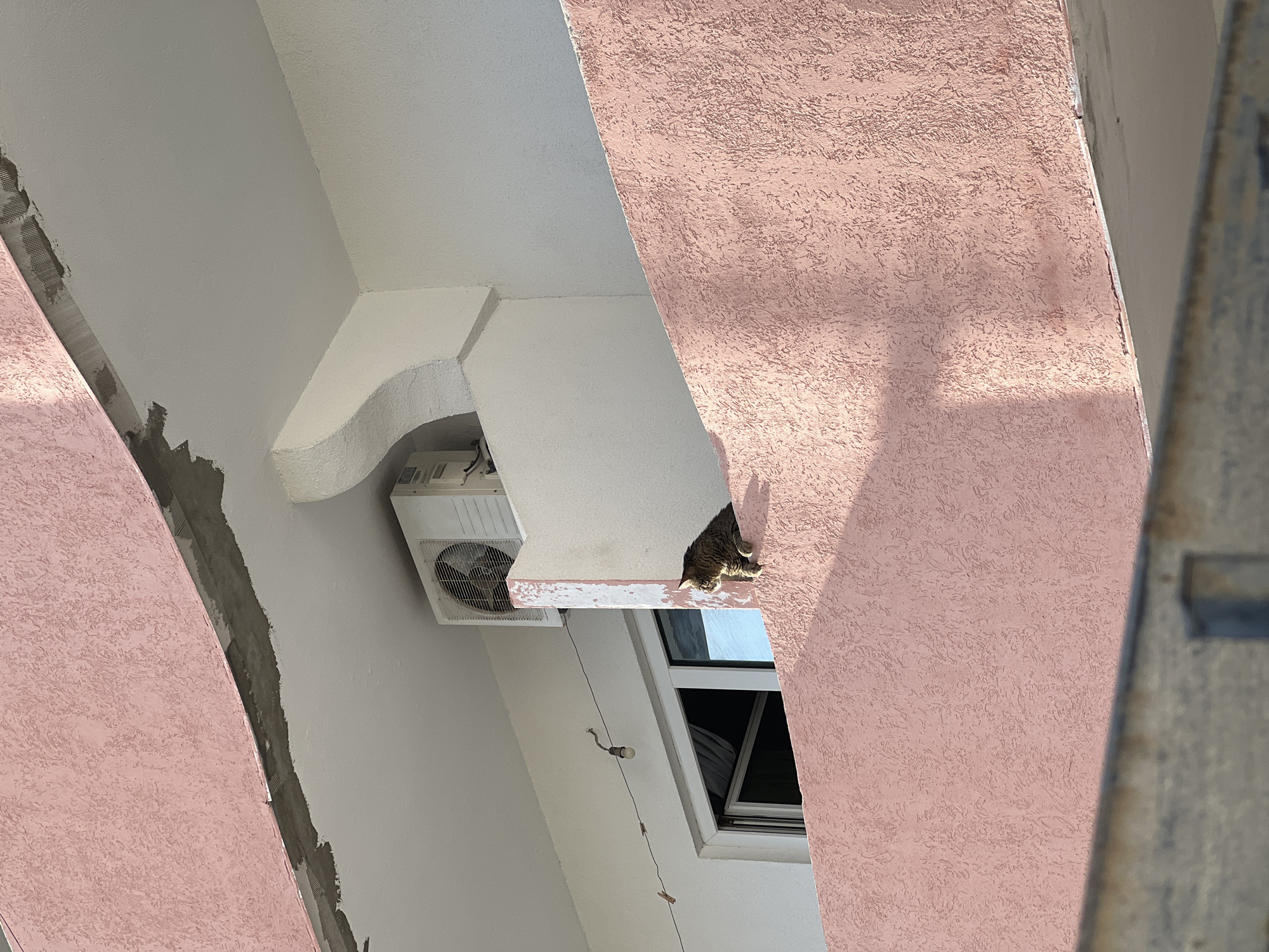 This media file presents an exterior view of a building in Budva, Montenegro, characterized by a mix of architectural styles and featuring a single tabby cat.
The scene is dominated by two large, pink-hued architectural elements, possibly columns or decorative panels, with a heavily textured, stucco-like finish. These contrast with smooth, white-painted walls and ceilings that form the background. Towards the upper right of the image, nestled on a white ledge above a partially open window, sits a tabby cat. Its striped and spotted fur is visible, and it appears to be resting or observing its surroundings, with part of its body in shadow due to the bright sunlight.
An outdoor air conditioning unit, light grey and white in color, is prominently mounted on a white wall to the left of the window, beneath a distinctively curved white architectural feature. Below the AC unit, a wire string with a single light bulb and what appear to be small clothespins hangs loosely. The window beneath the cat is a simple, rectangular design, with its top section slightly ajar. Along the left side of the image, one of the pink architectural elements shows signs of ongoing construction or renovation, with exposed grey plaster and mesh material.
The bright, direct sunlight suggests it is daytime, casting clear shadows across the textured pink surfaces and partially obscuring the cat. The overall impression is a quiet, sunlit corner of a building, typical of a Mediterranean urban environment. There are no people visible, and no discernible text is present in the image.