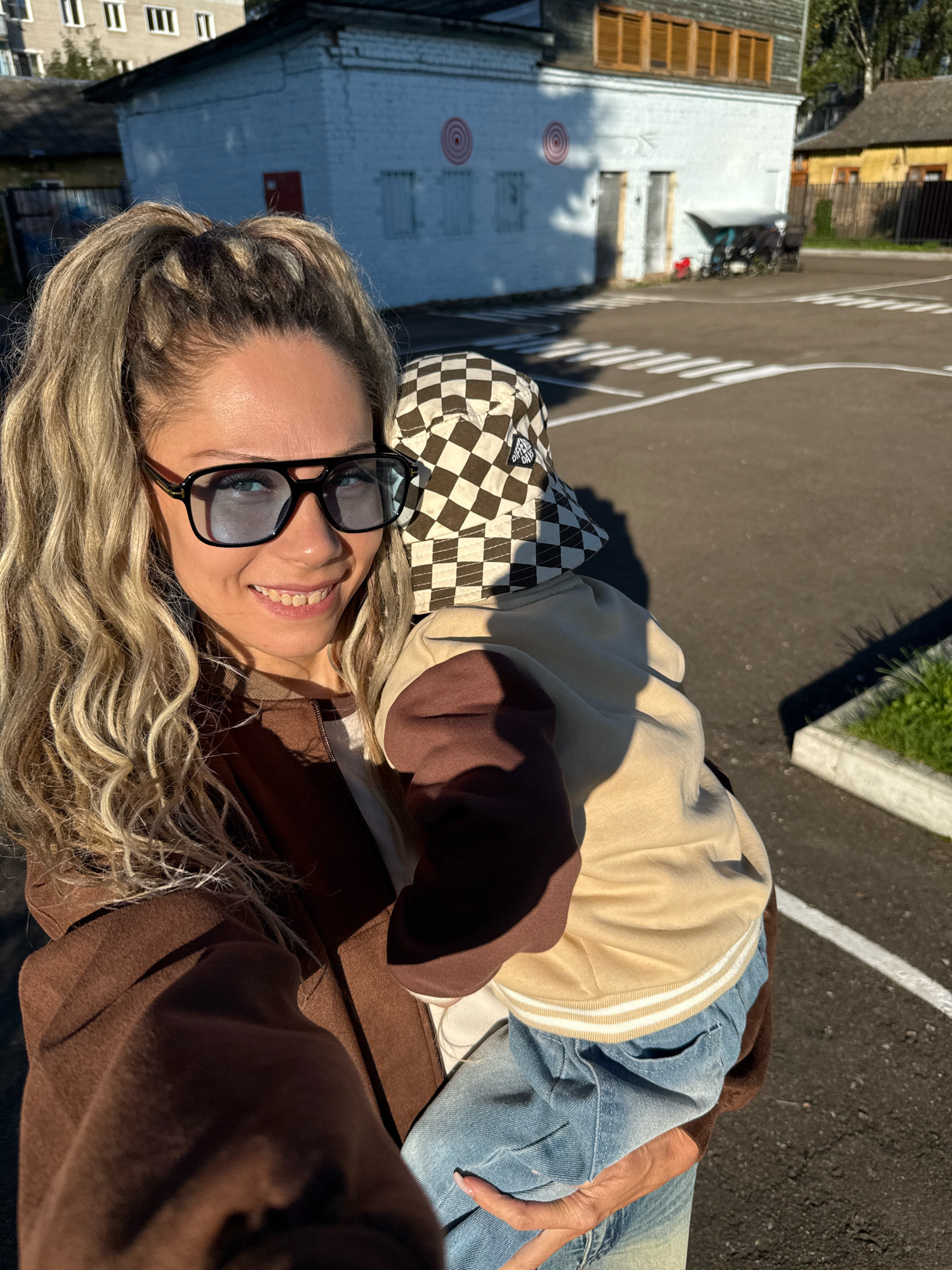 A medium shot captures a woman holding a baby outside a building in Rusanovo, Russia. The woman, positioned on the left, is looking at the camera and smiling. She has blonde, curly hair styled in a high ponytail and is wearing large, dark-framed glasses. Her attire consists of a brown coat over a white shirt and blue jeans.
The baby, held in the woman's arms, is facing away from the camera, partially obscured by the woman. The baby is wearing a beige jacket with brown sleeves, a white-striped collar, and a checkered bucket hat.
The background features a light-colored, brick building with several windows and two red circular targets on its exterior wall. To the right of the building, a yellow house with a dark roof is visible, along with a fence. The ground is asphalt, marked with white lines, suggesting an outdoor area or a parking lot. The scene is illuminated by the warm light of the sun, casting shadows.