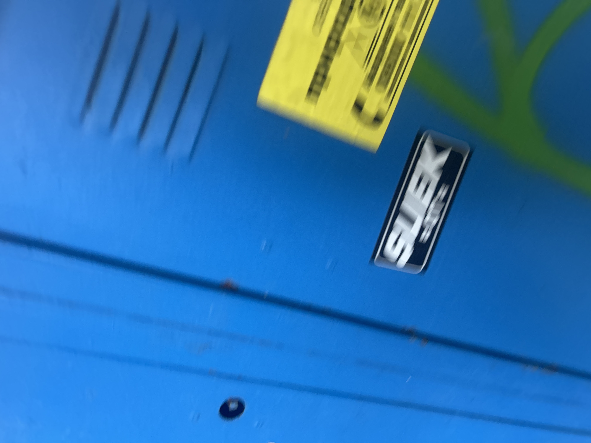 The image is a blurry, angled, close-up shot of a bright blue surface, possibly a locker or a wall. A rectangular black and silver sticker with the word "SLEK" is visible on the surface. To the right of the sticker, a partially visible yellow label with text is also present. The blue surface appears to have vertical lines, suggesting it might be made of multiple panels. A faint green graffiti-like marking is visible in the lower right quadrant of the image. The overall lighting suggests an outdoor setting. There are no people or animals in the frame.