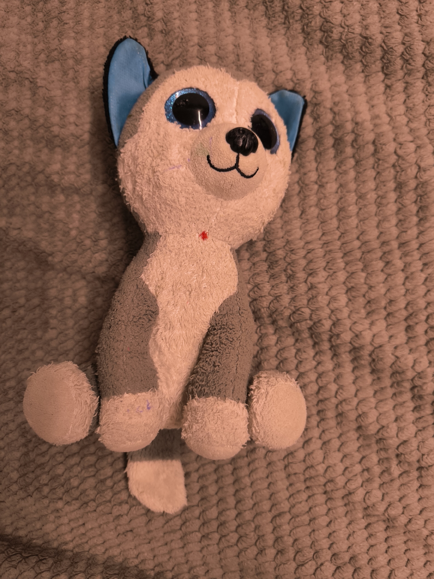 A plush toy dog sits upright on a textured, brown blanket. The toy has large, sparkly blue eyes, a black nose, and a small black smile. Its ears are blue with black tips, and its body is primarily white with grey patches on its back and legs. There is a small red embellishment on its chest. The toy is in focus, while the blanket behind it is slightly blurred. The image appears to have a sepia tone filter applied. There is no indication of the city of Октябрьский, Russia within the image.