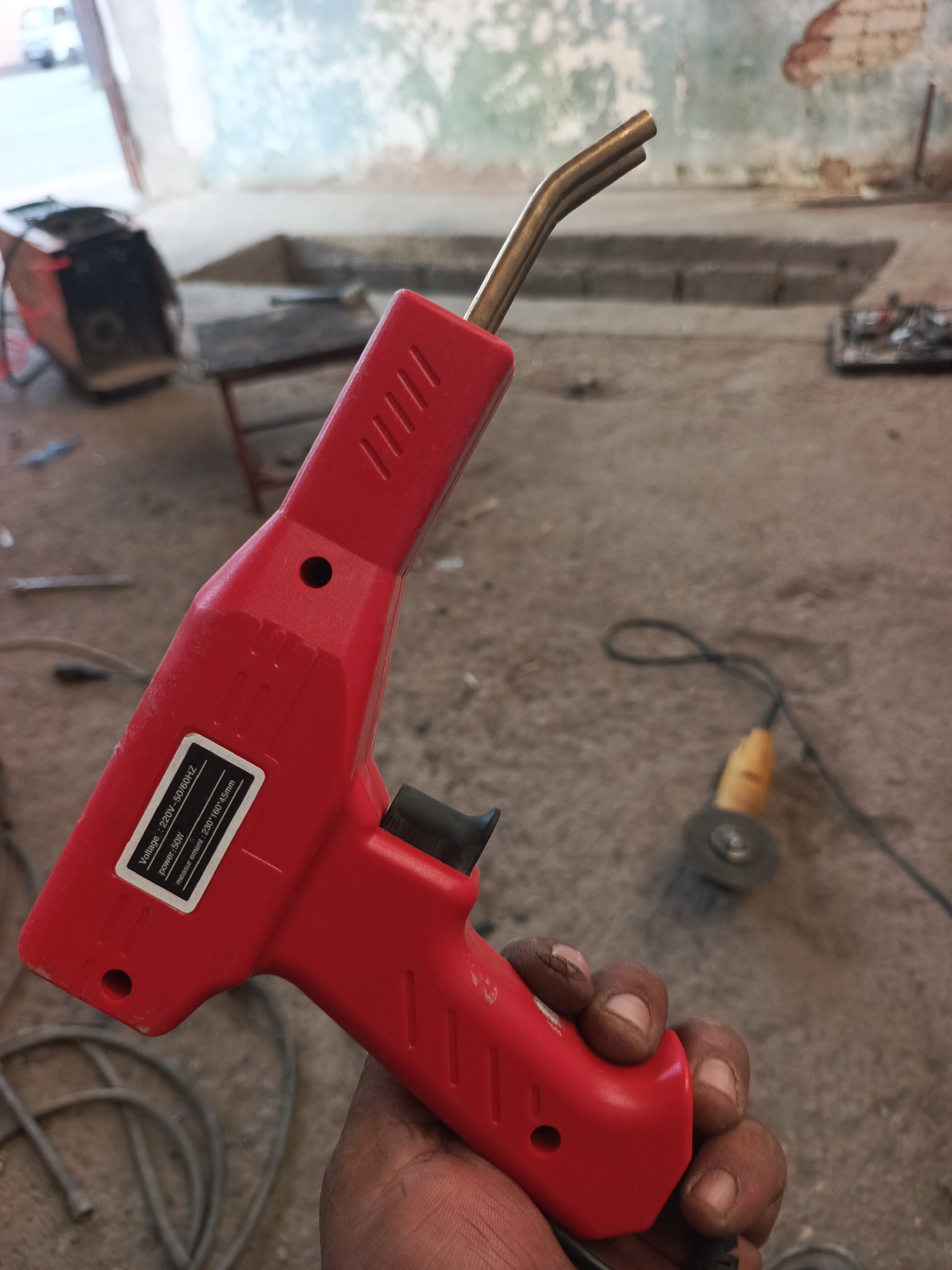 A close-up, eye-level shot shows a person's hand holding a red, heat gun with a metal nozzle that is slightly bent. The heat gun has a label that reads "Voltage: 220V - 50/60Hz", "power: 50W", and "measure ment: 230*160*45mm". The background is out of focus but appears to be an outdoor construction or workshop area, possibly in Kumkurgan, Uzbekistan, with a concrete floor, some rubble, and an out-of-focus power tool, possibly an angle grinder, in the middle ground.