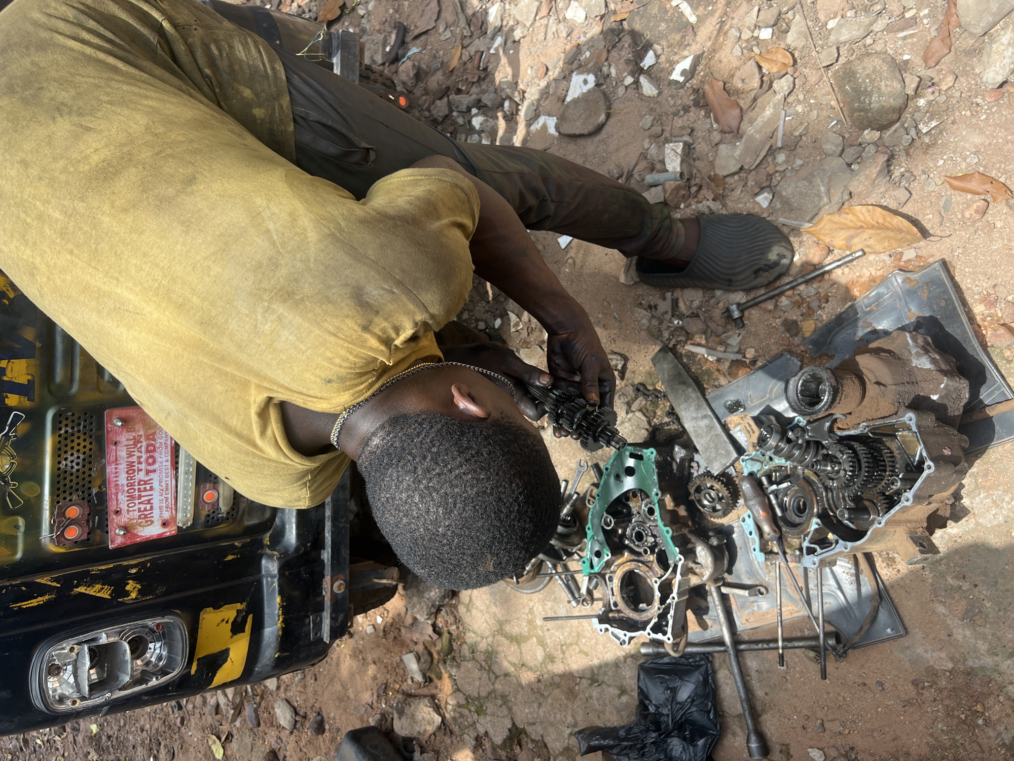 From an overhead perspective, a mechanic lies prone on the ground in what appears to be Nkpor, Nigeria, disassembling or reassembling a motorcycle or auto-rickshaw engine. The mechanic, an African male with a close-cropped hairstyle and dark skin, is wearing a yellow t-shirt, dark trousers, and grey slippers. His hands are covered in grease, and he is working with various engine components, including gears, shafts, and the engine casing.
These parts are laid out on the ground amidst debris, tools, and a black plastic sheet. Adjacent to the mechanic is a black vehicle or part of a vehicle with damaged lights and a sticker on it that reads "TOMORROW WILL GREATER THAN TODAY THIS IS MY PROMISE FROM GOD FROM EMMY BEST & COMPANY". The environment is predominantly composed of dirt, rocks, and scattered leaves, indicative of an outdoor workshop or repair location.