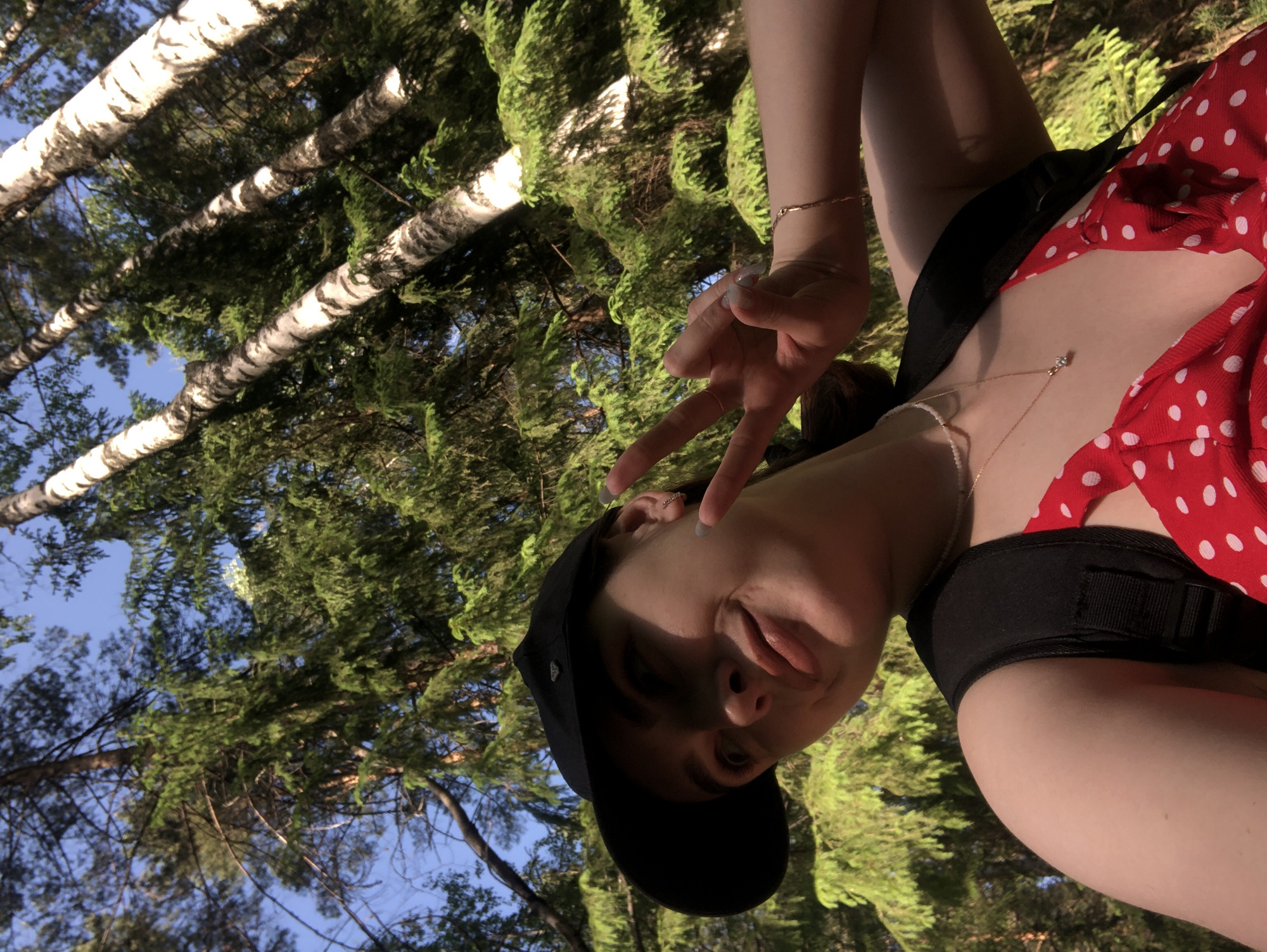 A young person, possibly female, with fair skin and dark hair wearing a black baseball cap, a red and white polka-dot top and a black tank top with thin gold necklaces and bracelets is shown from a low angle with trees visible in the background. The person is holding up their fingers making a peace sign. The location is potentially a forest or park in Noviy, Russia based on the presence of birch trees and pine trees in the background, both types of trees which are common in Russia.