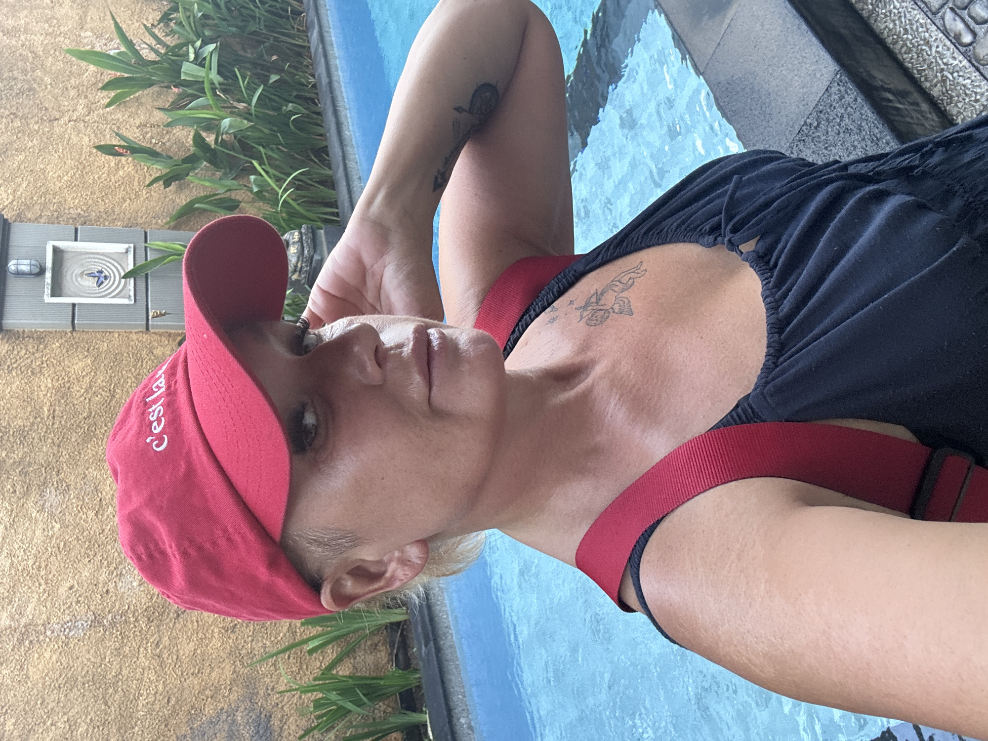 The media file shows a woman wearing a red baseball cap and a black top with red straps, posing next to a swimming pool. She has blonde hair and several tattoos, including one on her chest that appears to be of an angel or cherub, and another on her arm. The scene is outdoors, with lush green foliage visible in the background, and the water of the pool is a bright blue. The woman's expression is relaxed, and she appears to be enjoying a sunny day. The text "C'est la vie" is visible on the red cap. Based on the provided context, the location is Kuta, Indonesia. The overall mood is casual and leisurely, suggesting a vacation or a day of relaxation by the pool. The lighting indicates it is daytime, likely sunny.