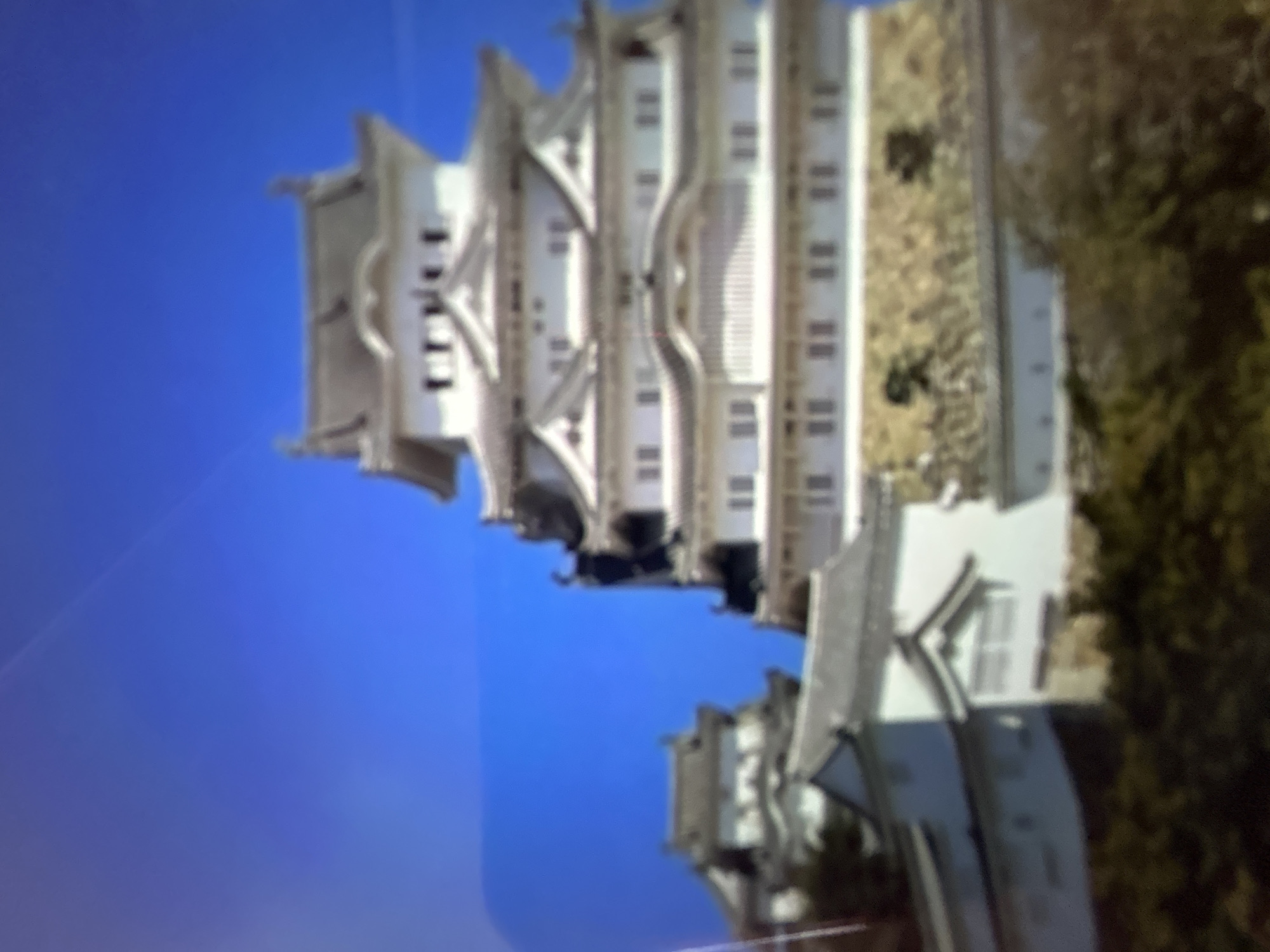 This media file captures a white, multi-tiered building with a traditional architectural style, reminiscent of a castle or historic monument. The building features numerous windows and curved rooflines. It is set against a clear blue sky, suggesting fair weather. The lower portion of the image shows a darker, possibly wooded area or foliage, and some additional smaller structures with white walls and dark roofs. The perspective is from a low angle, looking upwards at the main building. The image is slightly blurred, making precise details difficult to discern. There is no visible text in the image. Given the provided location context of Gelendzhik, Russia, this could be a depiction of a local landmark, though without further information or sharper detail, specific identification is not possible. The scene appears peaceful and offers a view of a significant architectural structure against a natural backdrop. The time of day is likely daytime, given the bright sky.