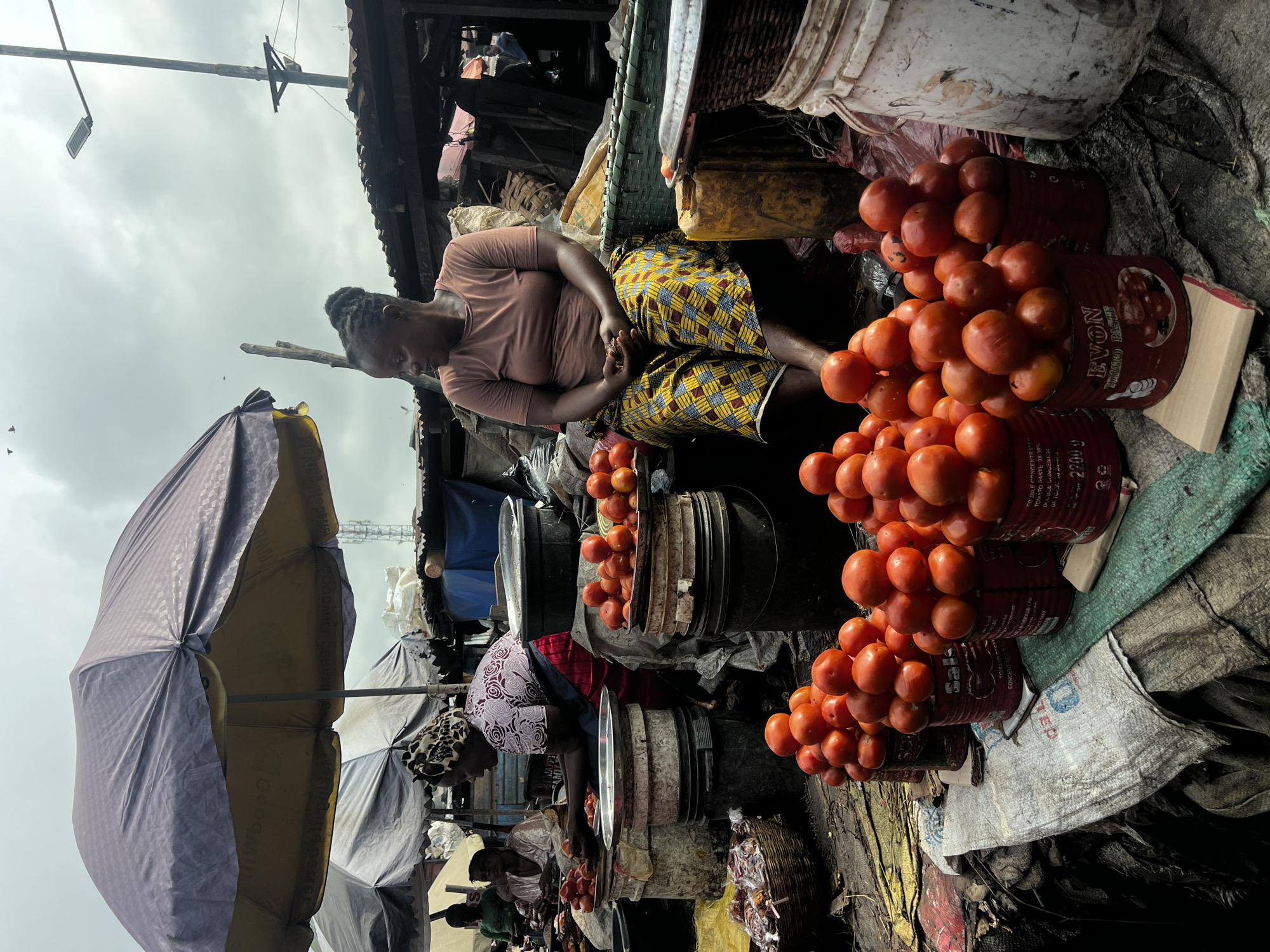At a bustling outdoor market in Nkpor, Nigeria, a woman is seated at a stall with a large quantity of tomatoes. She is wearing a light brown t-shirt and a colorful yellow and blue patterned skirt, with her hair tied up in a bun. Her hands are clasped in her lap as she looks to her left, away from the camera.
The stall is laden with produce, primarily tomatoes, which are piled high in various containers, including repurposed tin cans and buckets. In the foreground, several stacks of tomatoes are arranged in red, cylindrical cans labeled "EVON TOMATO PASTE." Behind her, more tomatoes are displayed on metal platters atop stacked buckets. A large, dark umbrella shades the stall from the overcast sky.
To the left of the main subject, another woman, wearing a patterned headscarf and a pink and white shirt, is attending to a pile of tomatoes in front of her. She is actively handling the produce, suggesting she is also a vendor or assistant. Other market stalls and structures, made of corrugated metal and wood, are visible in the background, indicating a busy commercial area. The ground is covered with sacks and assorted debris, characteristic of an open-air market. The overall scene captures a moment of daily commerce and activity in this Nigerian city.
