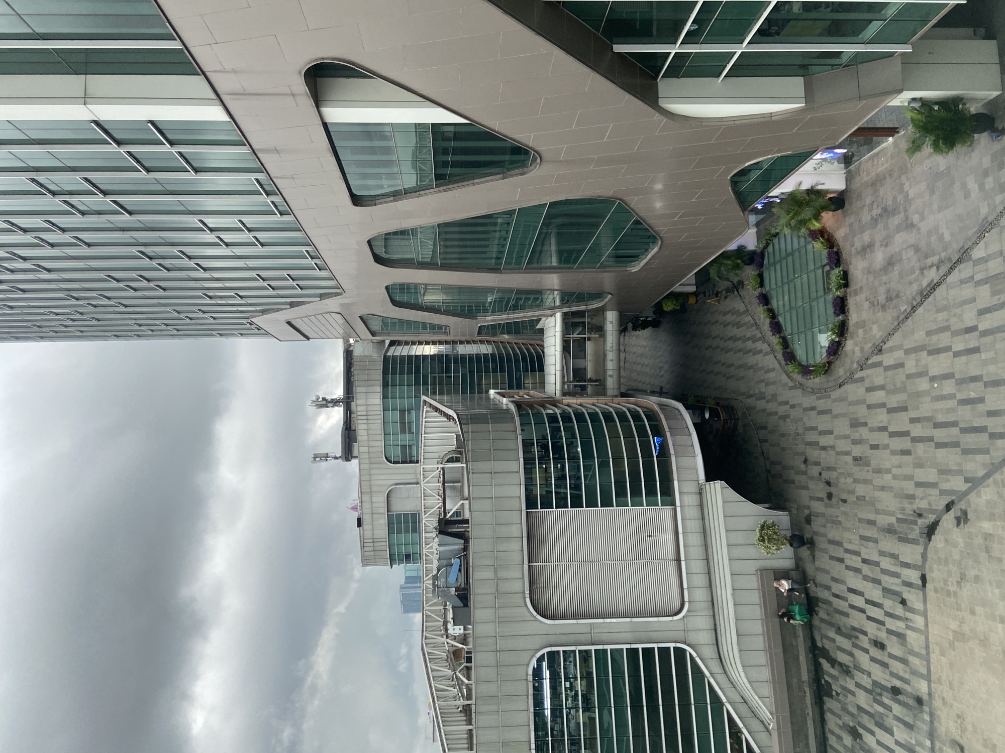 The image shows a partial aerial view of buildings in North Jakarta, Indonesia, under a cloudy sky. The architecture is modern, featuring a combination of glass and light-colored, possibly concrete or stone, facades. One building has distinctive cutout shapes in its overhanging structure. At ground level, there is a landscaped area with vegetation and paved surfaces. People are visible near one of the buildings. There is a bench where two people may be sitting. The photograph is taken at an angle, making it appear as if the view is rotated.