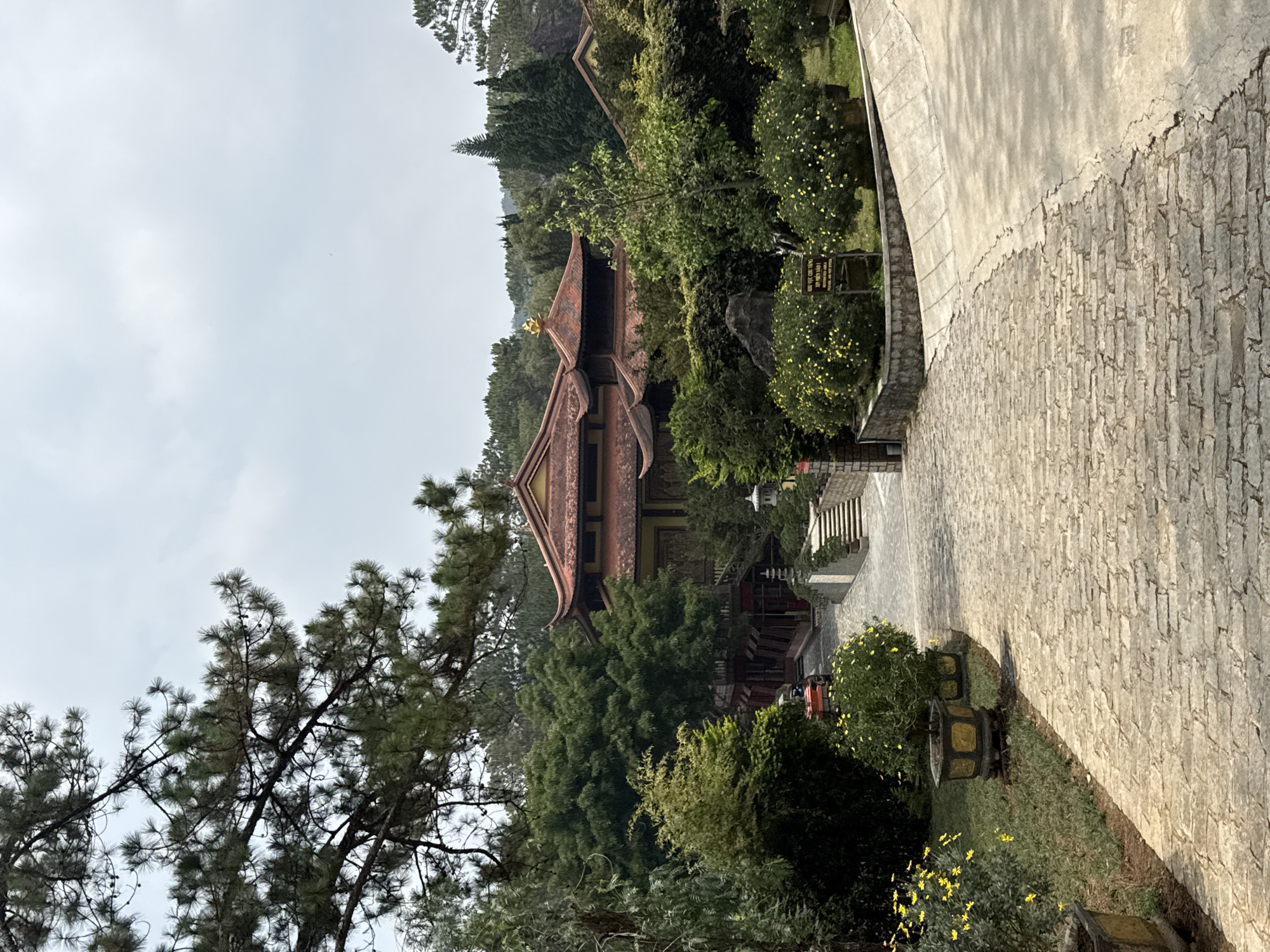 The image shows the Truc Lam Zen Monastery in Da Lat, Vietnam. The view is of the monastery building with a red-tiled roof and golden ornamentation, surrounded by green trees. A stone pathway leads towards the building, with plants in decorative pots lining the path. The sky is overcast. There are no people visible in the frame.