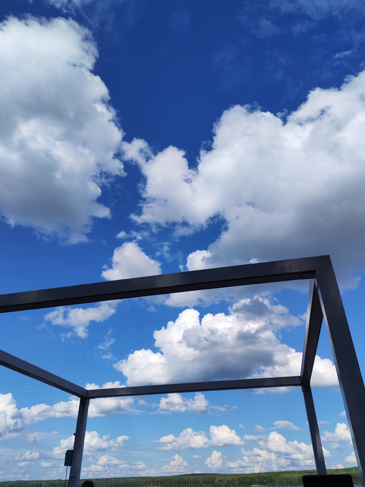The image captures a view of a bright blue sky filled with fluffy white and grey clouds. In the foreground, a dark gray metal structure, forming a corner of a geometric frame, bisects the sky. This structure appears to be part of a larger architectural element, possibly a canopy or a balcony railing, viewed from a low angle.
Below the sky and the metal structure, a distant landscape is visible. It features a dense line of green trees, suggesting a forest or a heavily wooded area, stretching across the horizon. A sliver of what might be water or a very flat plain is also discernible at the very bottom edge of the image. The overall impression is one of an outdoor setting on a clear, cloudy day. The lighting suggests it is daytime, likely late morning or afternoon, given the brightness of the sky and the distinct shadows cast by the clouds. There are no discernible emotions evoked, and no visible text. The context provided places this scene in Chaikovsky, Russia.