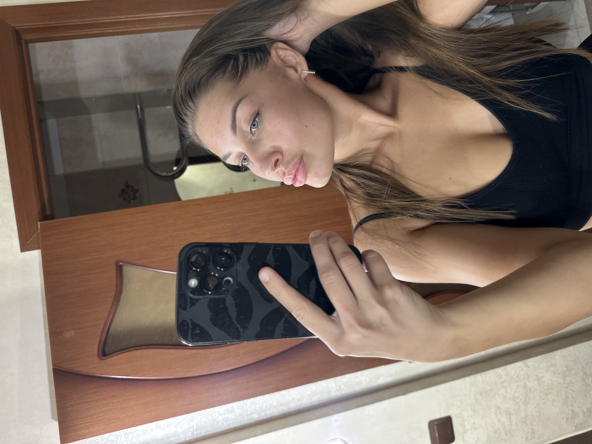 A young woman is captured in a selfie taken in Irkutsk, Russia, holding her smartphone to photograph her reflection. The image shows her from the chest up, her light brown hair falling over her shoulders and pulled back behind her left ear, revealing a small hoop earring. She has fair skin, light blue eyes, and full, light-colored lips, forming a pouty expression. She is wearing a simple black ribbed top or sports bra.
Her left hand holds a dark-colored iPhone, which has a distinct case featuring a pattern of dark lip prints or kisses. The camera module of the iPhone, with its multiple lenses, is clearly visible. Her right arm is bent, with her hand resting on the back of her head, lightly touching her hair.
The setting appears to be indoors, likely a room with wooden trim around what seems to be a mirror. The reflection in the background shows a light-colored tiled wall, possibly indicating a bathroom or dressing area. Below the phone, a wooden surface with a shiny, wavy, golden-brown decorative inlay is visible. The lighting suggests an indoor environment with artificial light. No visible text is present in the image.