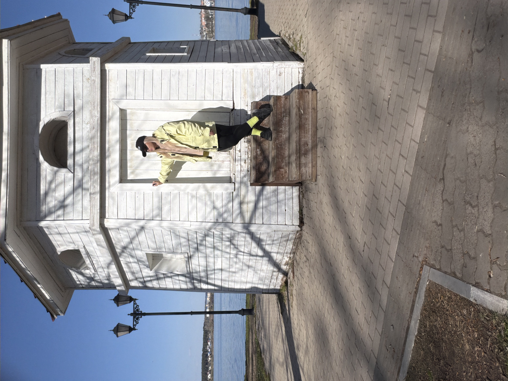 The image shows a person wearing a yellow jacket, black pants, and a black cap standing on a wooden bench outside a small white building. The building has a unique window shape on the side and is located near a body of water, as indicated by the presence of the water visible in the background. Black lampposts stand beside a stone walkway. The location is identified as Votkinsk, Russia.
